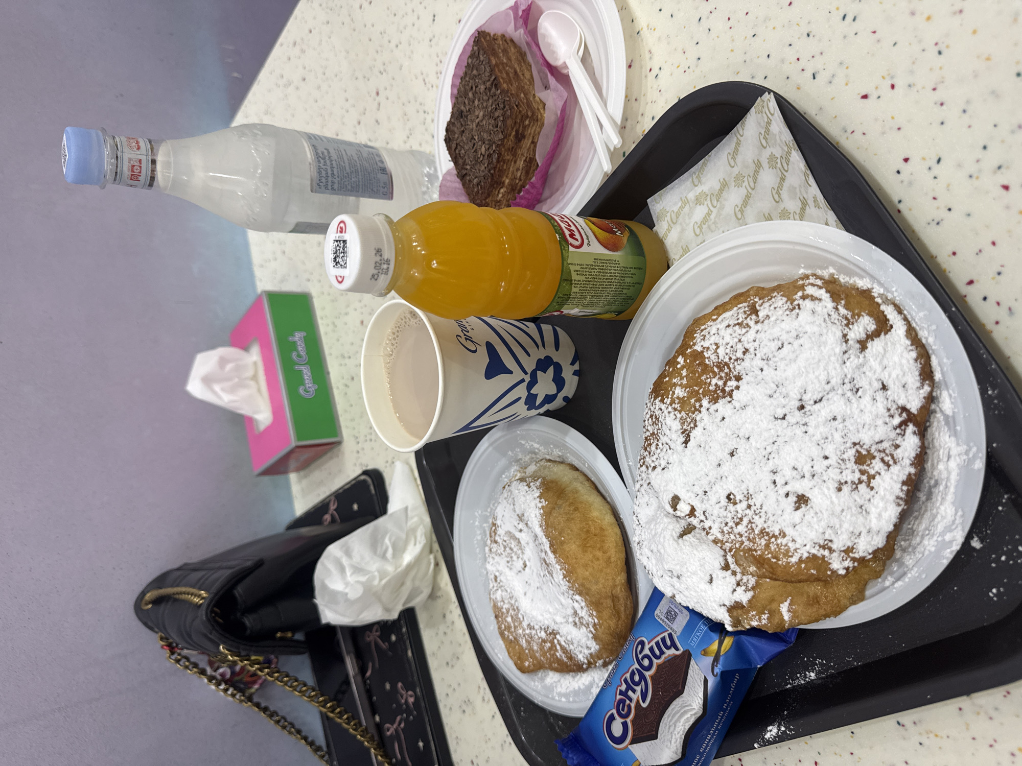 A meal or snack is displayed on a black tray on a light-colored, speckled countertop. The scene is likely indoors, possibly in a cafe or food court in Yerevan, Armenia.
On the tray, there are two large, golden-brown pastries heavily dusted with powdered sugar, served on white disposable plates. Next to one pastry is a packaged ice cream sandwich. Another white disposable plate holds a piece of cake with dark chocolate shavings. Drinks include a clear bottle of still water, an orange-colored juice bottle, and a paper cup containing a light-colored beverage, possibly coffee with milk.
Other items visible around the tray include a pink and green box of "Grand Candy" tissues and a black purse with a gold chain.
Visible text includes:
* "Grand Candy" on the tissue box, paper cup, and some wrappers.
* "Сендвич" (Cyrillic for "Sandwich") on the blue ice cream package, along with "МЯГКОЕ П", "шоколадным печеньем", and "ное ванильный пломбир" (indicating soft chocolate cookies and vanilla ice cream).
* "Ma" on the juice bottle, and a date "25.02.26 10.16" on its cap.
* The water bottle label reads "Քյուրեղ" աղբյուր բնական խմելու ջուր չգազավորված (Armenian for "Kyuregh spring natural drinking water uncarbonated") and "0.5л".
QR codes are present on the water bottle cap and the ice cream wrapper.