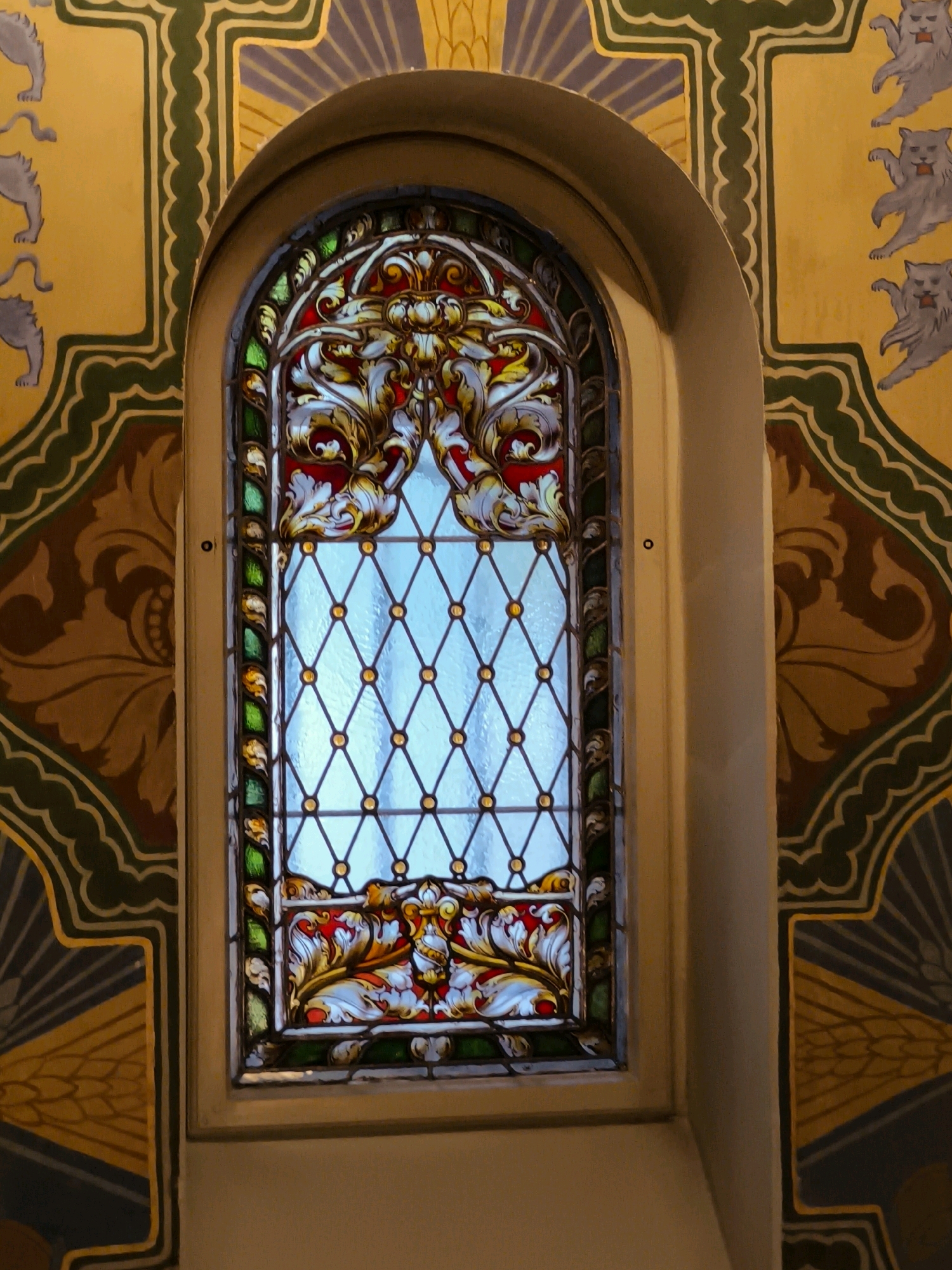 The image shows a close-up view of a stained-glass window framed by ornate, decorative wall paintings. The stained-glass window is arched and features a geometric diamond pattern in the upper section, with colorful floral motifs in red, white, gold, and green in the lower section. The surrounding walls are painted in yellow with dark green borders and elaborate patterns, including stylized cat figures and floral elements. The lighting suggests an interior setting, possibly a historical building. The scene is devoid of any people or explicit actions. The architectural style and decorative elements suggest a European context, and given the prompt, the location is specified as Tallinn, Estonia, likely within a church or a historically significant building.