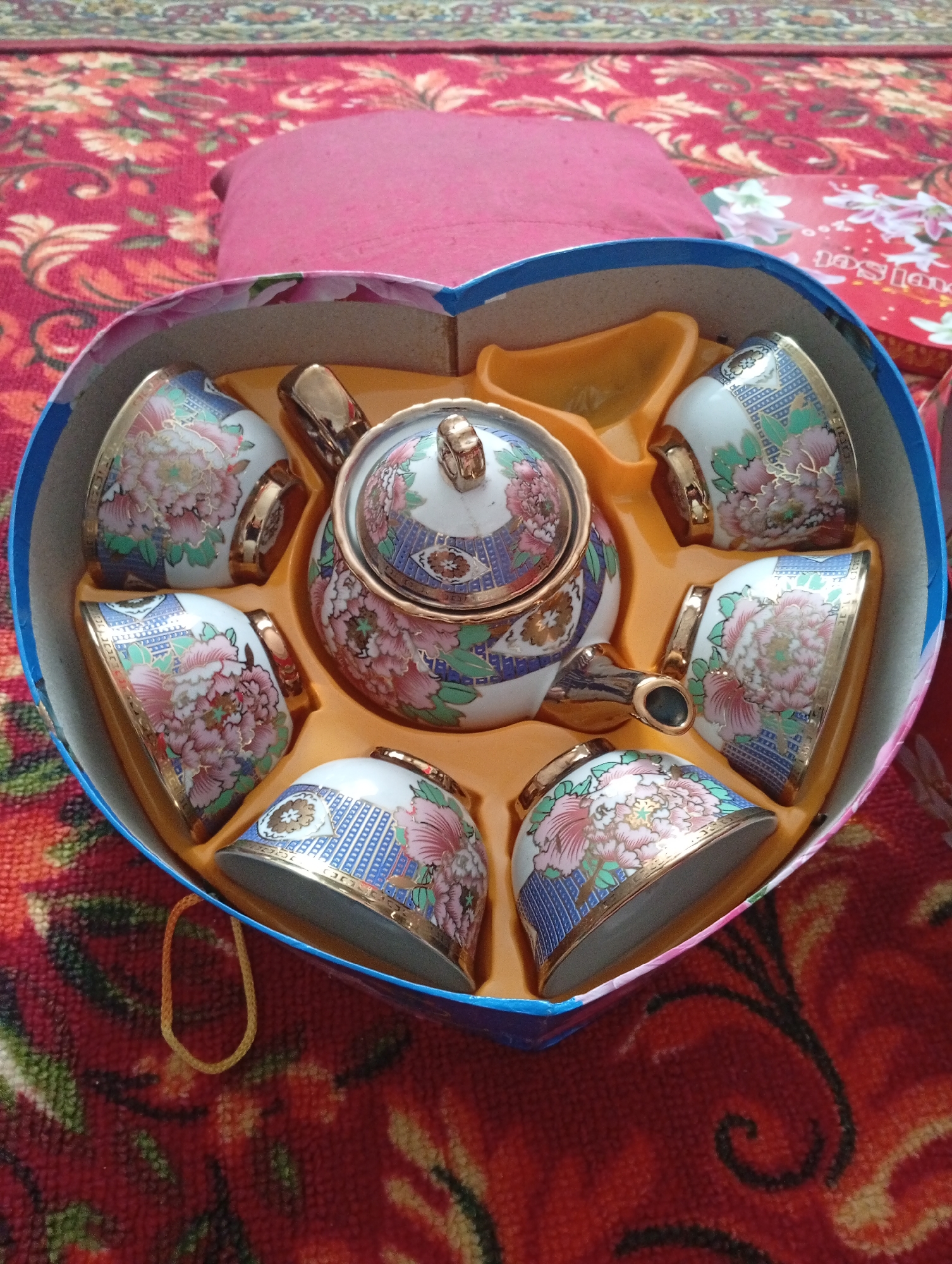 A heart-shaped gift box containing a tea set sits on a red, patterned fabric. The tea set includes a teapot with a lid and five cups. Each piece is decorated with floral patterns in pink, green, and blue, and trimmed with gold. The interior of the box is lined with orange plastic to hold the pieces in place. The background fabric has an intricate red, orange, and yellow floral design. The scene is indoors.