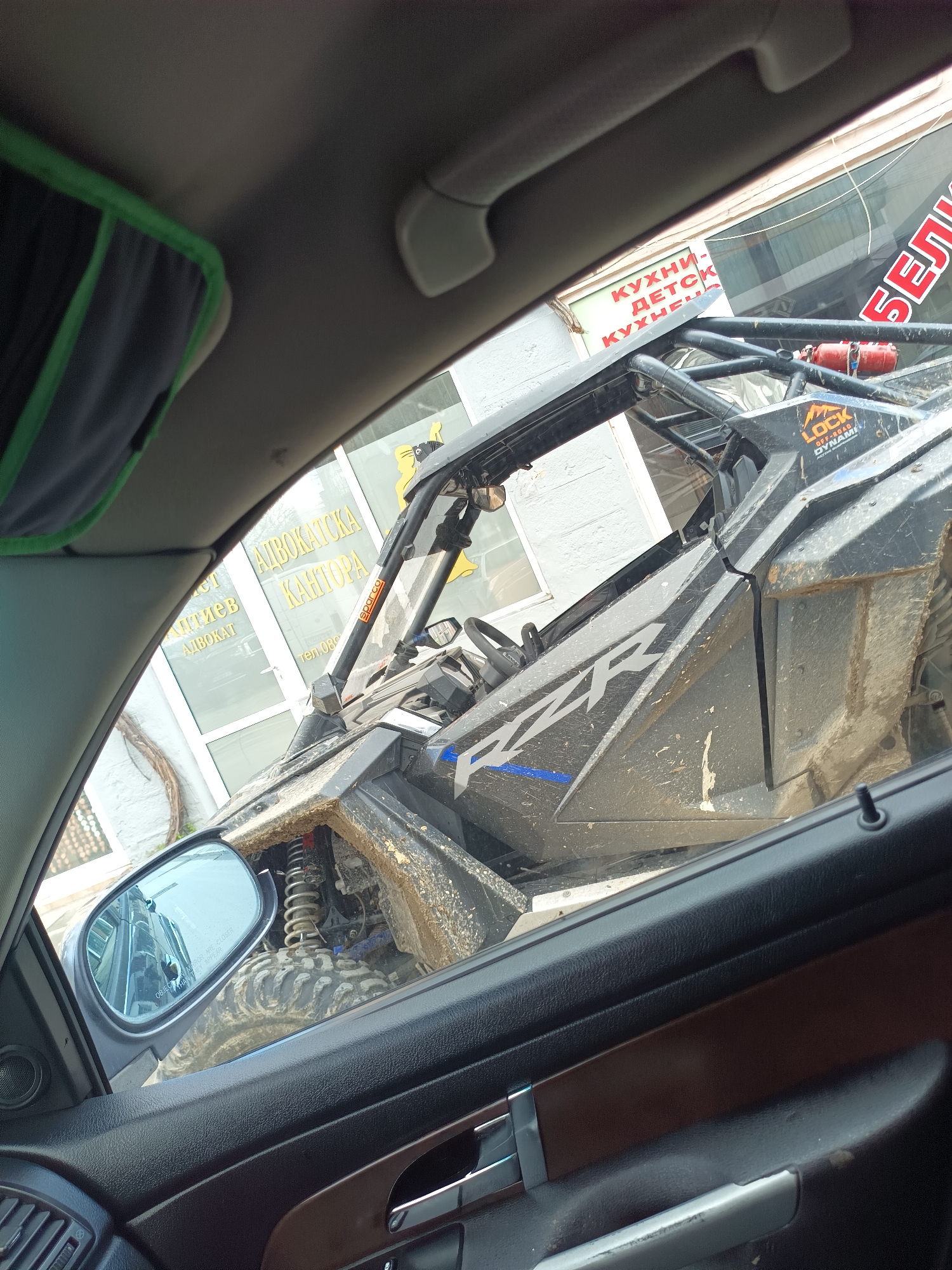 The image, taken from inside a car, shows a muddy Polaris RZR off-road vehicle parked outside of buildings in Targovishte, Bulgaria. Signs for "Advokatska Kantora" (Law Office) and "Kuhni Detski Kuhni" (Kitchens Children's Kitchens) are visible, along with the first few letters of "BELI" (White). The car door's interior, including the door handle and side mirror, is in the foreground.