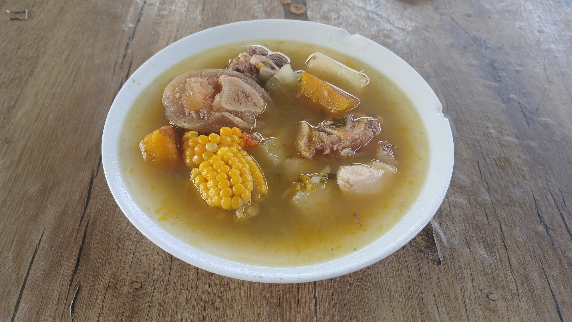 A white bowl of soup is centered on a wooden table with a repeating wood grain pattern. The soup contains large pieces of meat, a section of corn on the cob, diced pumpkin, and sections of cassava root. The bowl has a slight chip on its right side. The soup's broth is a pale yellow color, with some visible herbs and spices. No people are visible. The image was likely taken in Altagracia, Venezuela.