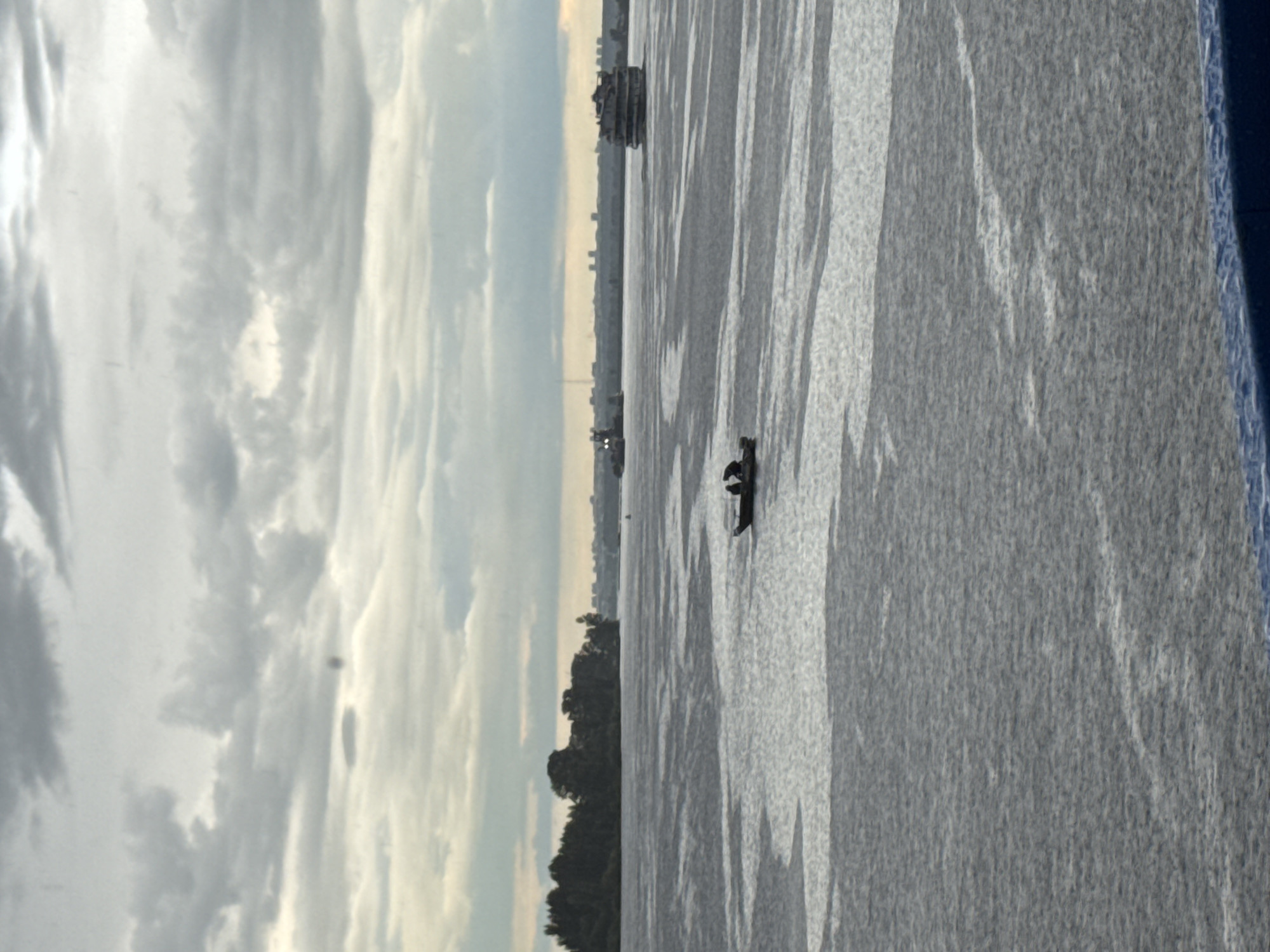 The image depicts a wide, gray river under an overcast sky with scattered clouds. A dark green tree line runs along the left bank, with buildings visible in the distance on the right bank. In the foreground, a small, dark boat with two occupants is navigating the rippling water. Further out on the river, a larger, multi-decked vessel, likely a passenger ship, is visible, as is another, darker vessel in the distance to the left. The scene suggests a river port or waterway in the city of Yelabuga, Russia, possibly during daylight hours.