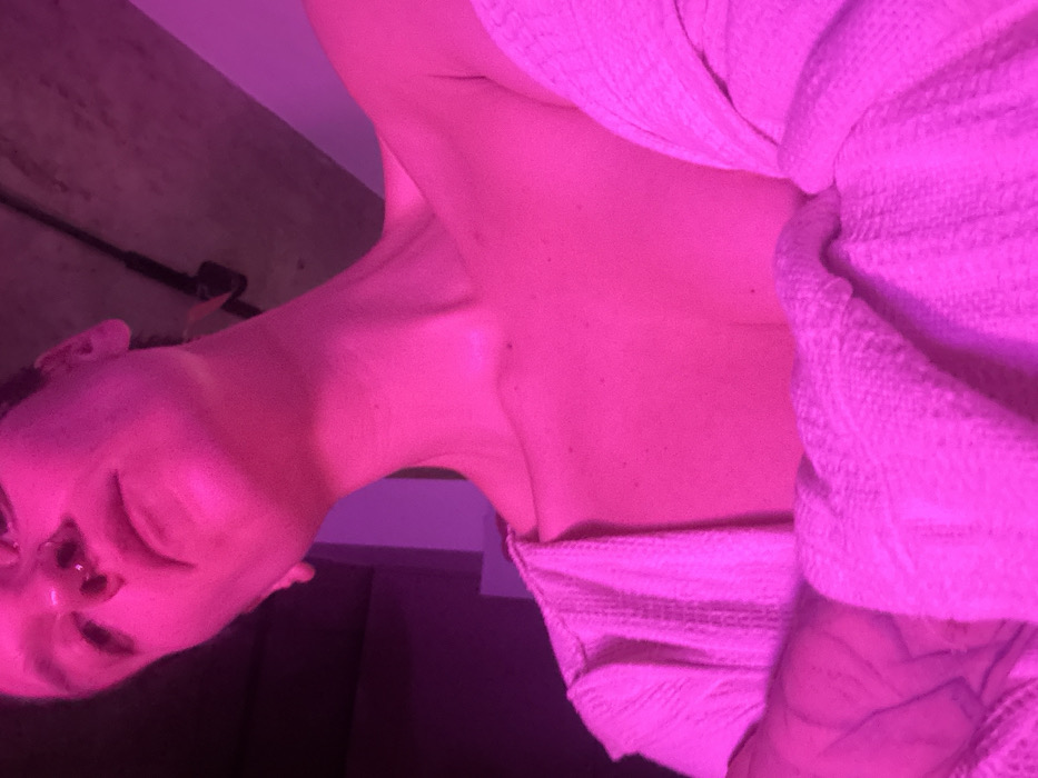 A person is wrapped in a textured, light-colored fabric or towel. The skin appears pale and speckled. The lighting is predominately pink, casting a magenta hue over everything. The person's face is partially visible, with closed eyes and a nostril piercing evident. The angle is tilted, with a ceiling and a dark wall appearing in the background. The composition is centered on the person's upper body, highlighting the texture of the fabric and the details of the skin.