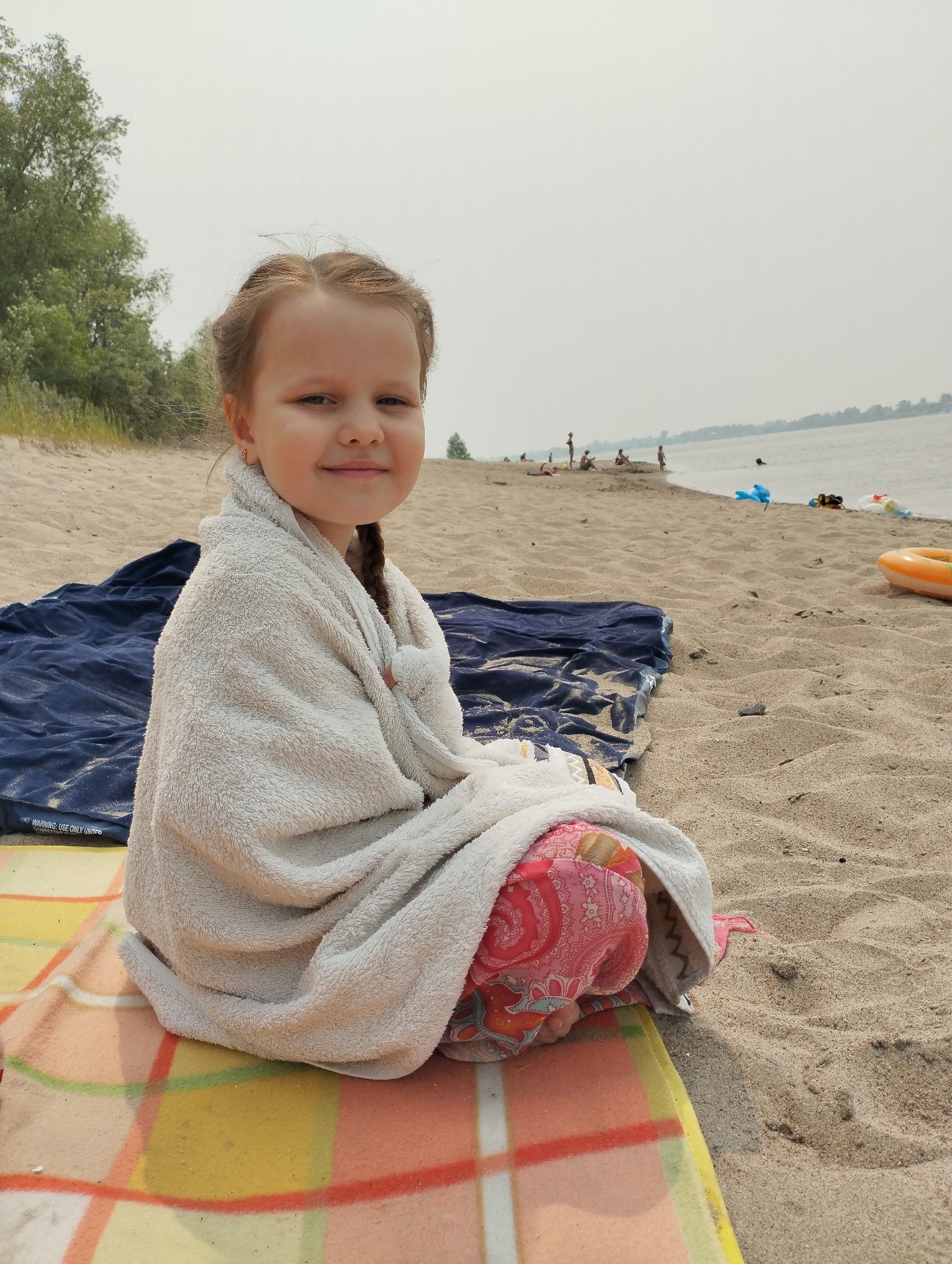 A young girl with braided brown hair, wrapped in a light-colored towel, sits on a beach. She is wearing colorful patterned pants underneath the towel and has a small gold earring. The girl is smiling at the camera. She is seated on a striped blanket laid out on the sand, with a dark blue towel behind her. In the background, there is a body of water, likely a river or lake, with a hazy sky and trees lining the distant shore. Several other people are visible in the distance, some appearing to be swimming or relaxing on the beach. There are also some colorful beach items scattered on the sand, including an orange inflatable ring. The scene appears to be set on a sunny but hazy day. The visible text on the blue towel reads "WARNING: USE ONLY UNITED".