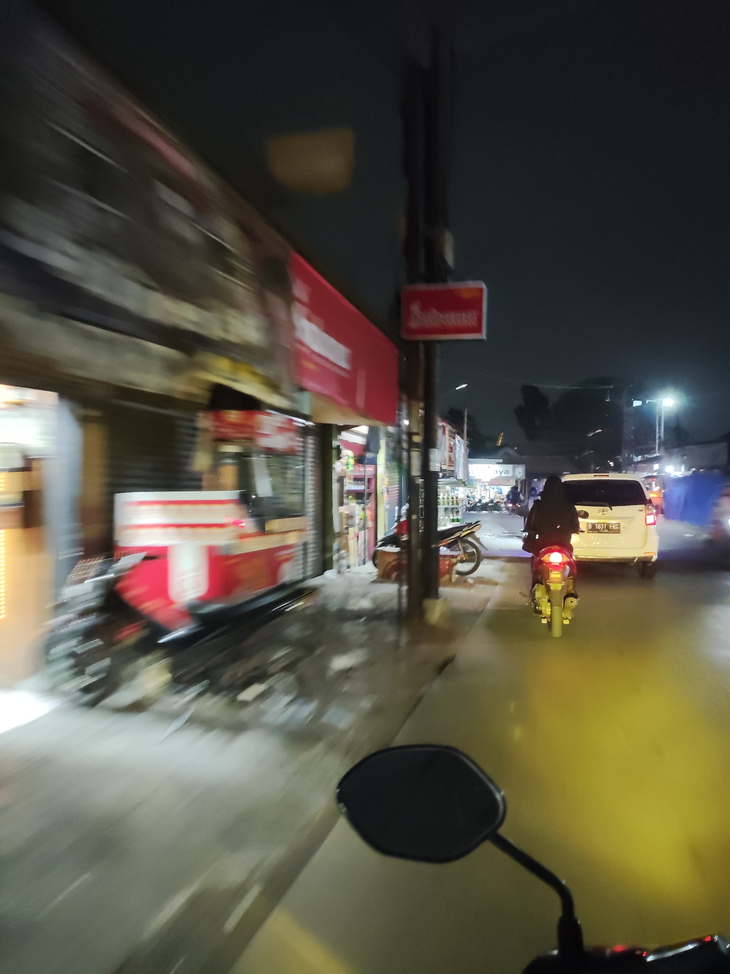 The image depicts a nighttime scene on a street in Telagaasih, Indonesia. A person is riding a motorcycle, with a portion of the motorcycle's handlebar and mirror visible in the foreground. The rider is wearing dark clothing and a black headscarf. Ahead of the motorcycle, a white car is driving on the road, with its license plate reading "B 1837 FRR". On the left side of the street, there are several buildings with illuminated signs, including a red sign with white text and a storefront that appears to be a food stall or shop with a red and white striped cart. A parked motorcycle is also visible on the left. The street is wet, reflecting the lights from the surrounding environment. The overall atmosphere is that of a busy urban street at night.