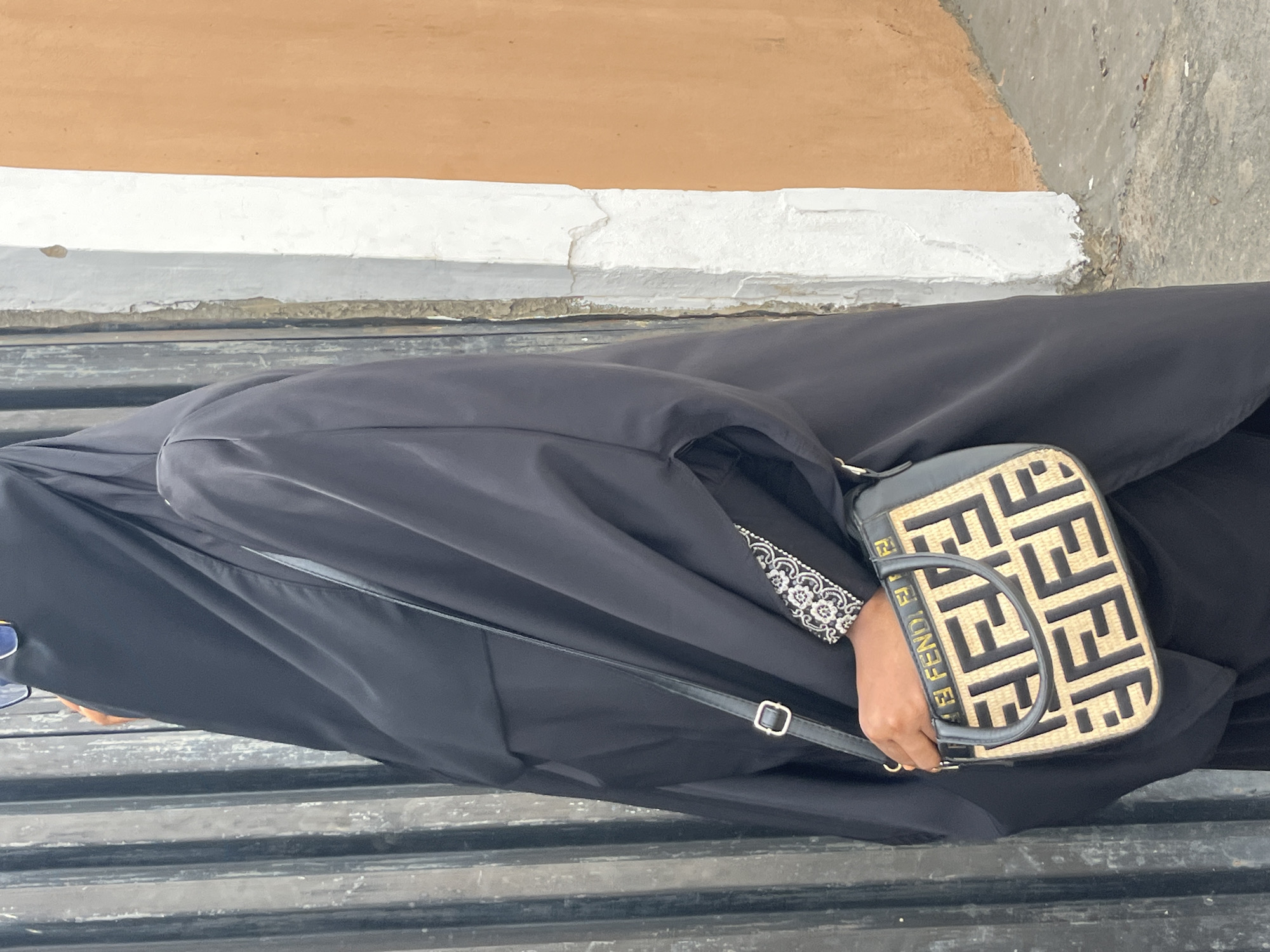 An overhead, slightly angled shot shows a person, likely a woman, seated in what appears to be an outdoor or semi-outdoor setting. The person is dressed in a flowing, black abaya or similar garment, which covers most of their body. A glimpse of patterned fabric, possibly an embroidered or embellished cuff in white and black, is visible on their right sleeve.
The person is holding a small, rectangular handbag with a distinct design. The handbag features a light, possibly straw or woven, texture base with a prominent repeating pattern of interlocking black "FF" logos, a signature design element. The strap and trim of the bag are black leather. On the front of the bag, the brand name "FENDI" is also visible in black letters on a lighter background, and it appears again on the strap. A metal buckle is seen on the bag's shoulder strap.
The person is seated on a dark, slatted surface, which could be a bench, fence, or railing. In the background, there's a wall or structure with distinct horizontal sections. The uppermost section is painted a light brown or tan color. Below this is a horizontal band of white, which shows some texture and cracking. Below the white band, there's a rougher, greyish surface, possibly concrete or stone. The lighting suggests it is daytime. The provided context indicates this image was taken in Ado, Nigeria.