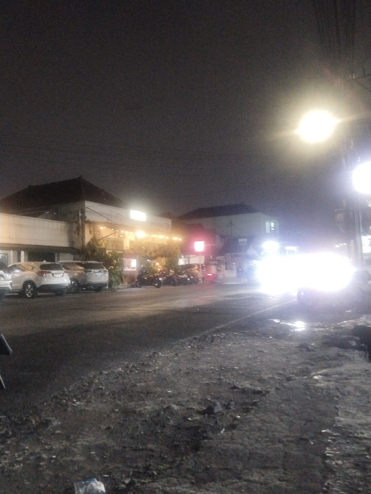 A nighttime street scene in Unknown, Unknown, features parked cars and motorcycles in front of a lit building. The street is dark and paved, with some debris. A bright streetlight illuminates the scene. The building appears to be a restaurant or commercial establishment with outdoor seating and string lights. Several cars, including SUVs, are parked parallel to the building. Motorcycles are also parked in front of the building. The sky is dark, suggesting nighttime.