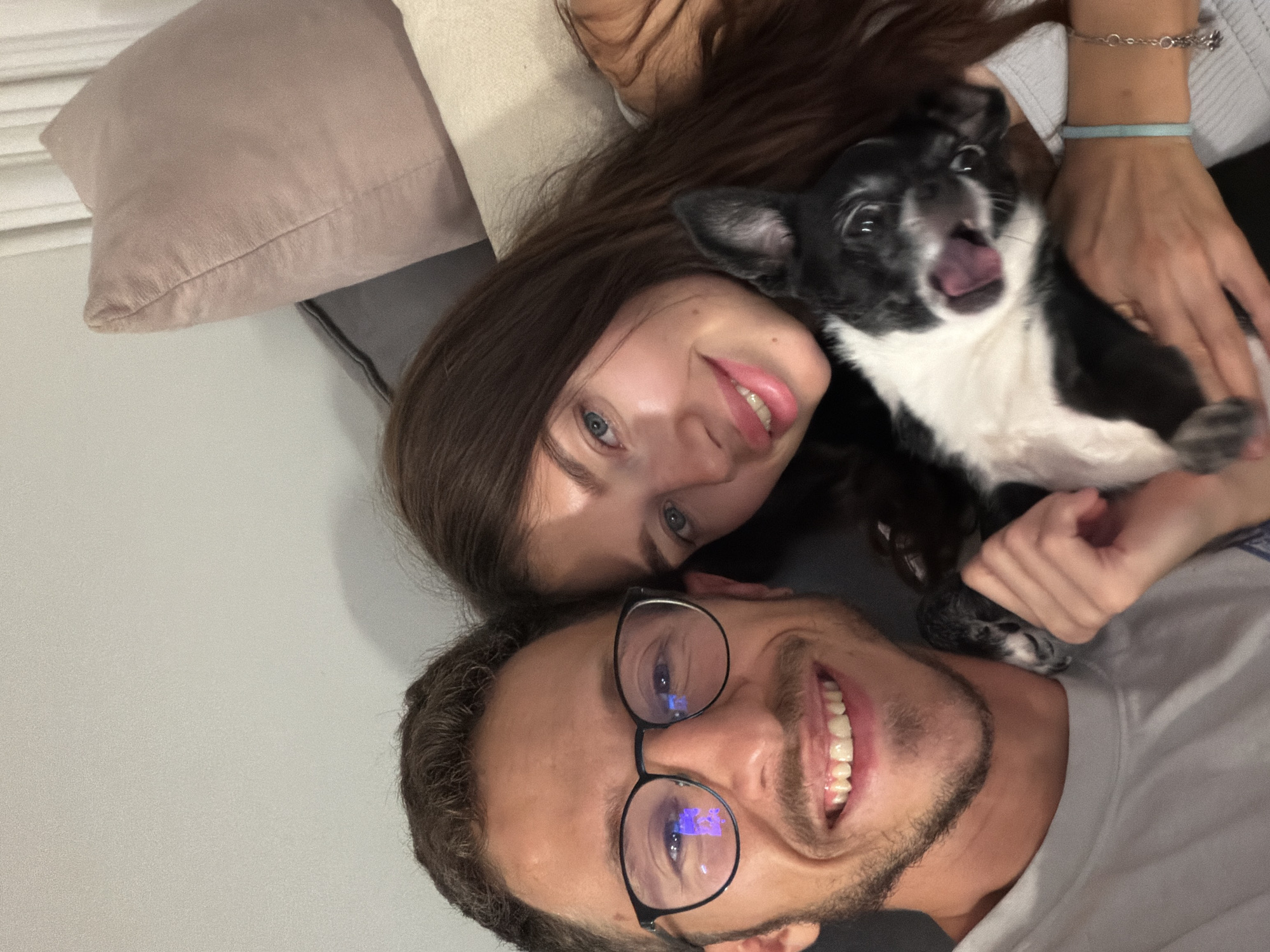 A selfie-style photograph shows two people and a small dog. A woman with long, dark hair and blue eyes smiles at the camera, while a man wearing glasses is positioned beneath her, also smiling. The small, black and white dog is held by the woman, its tongue sticking out. They appear to be indoors on a couch with cushions. There is no clear indication of the location being Tel-Aviv, Israel.