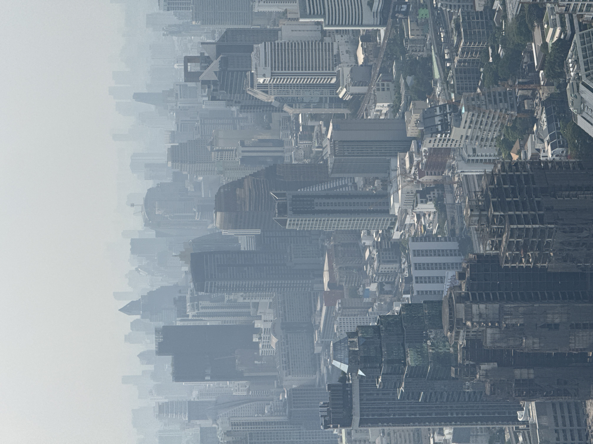 This is an aerial view of the city of Bangkok, Thailand. The image shows a dense cityscape with numerous skyscrapers and buildings, many of which are partially obscured by haze or pollution. The buildings vary in design, with some appearing older and more traditional, while others have a modern, glass-clad aesthetic. Construction cranes are visible, indicating ongoing development in the area. A network of roads and possibly elevated rail lines can be discerned among the buildings. The overall impression is of a bustling, densely populated urban environment experiencing air quality issues.