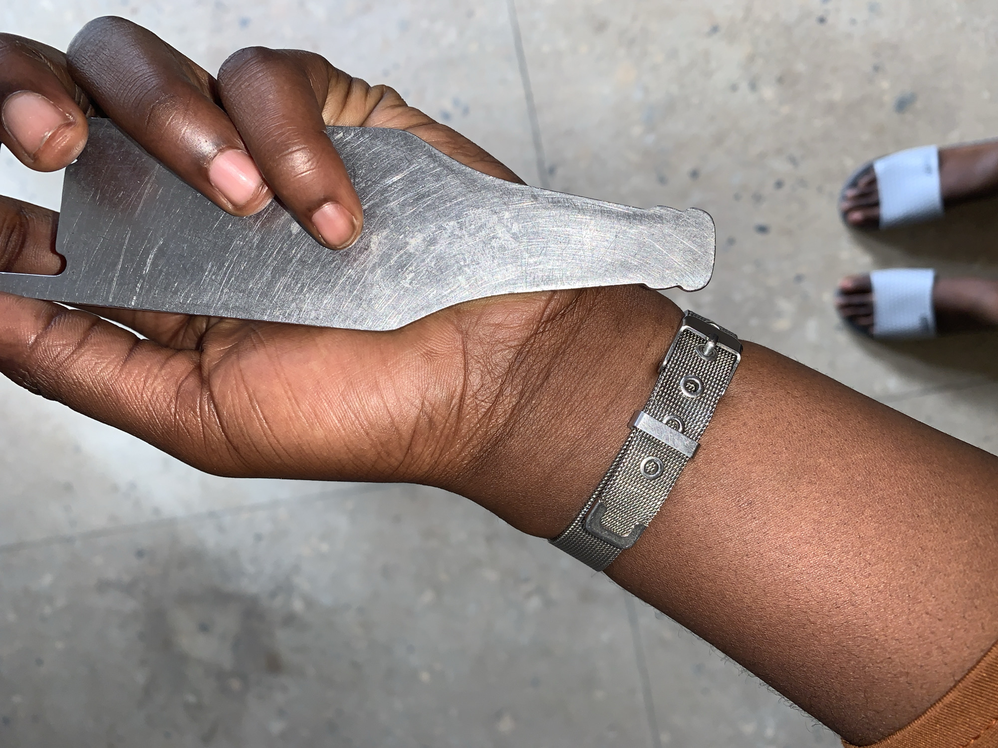 A close-up, high-angle view in the city of New Stadium, Nigeria, captures the left arm and hand of an individual with dark skin, holding a metallic object, with their feet visible on a concrete surface. The forearm, extending from the bottom left, wears a silver-toned mesh wristband with a buckle closure and several small circular rivets. The hand firmly grips an elongated, flat metallic object characterized by a brushed or scratched surface with numerous parallel striations. The object's top edge features a distinct rectangular notch, and its bottom tapers slightly. The fingernails on the visible fingers (thumb, index, middle, ring, pinky) are short and appear to have a clear or light pink polish. Below the hand, two feet, consistent with the arm's skin tone, are visible wearing light-colored slip-on sandals. The background consists of a light gray, speckled concrete or tiled floor.