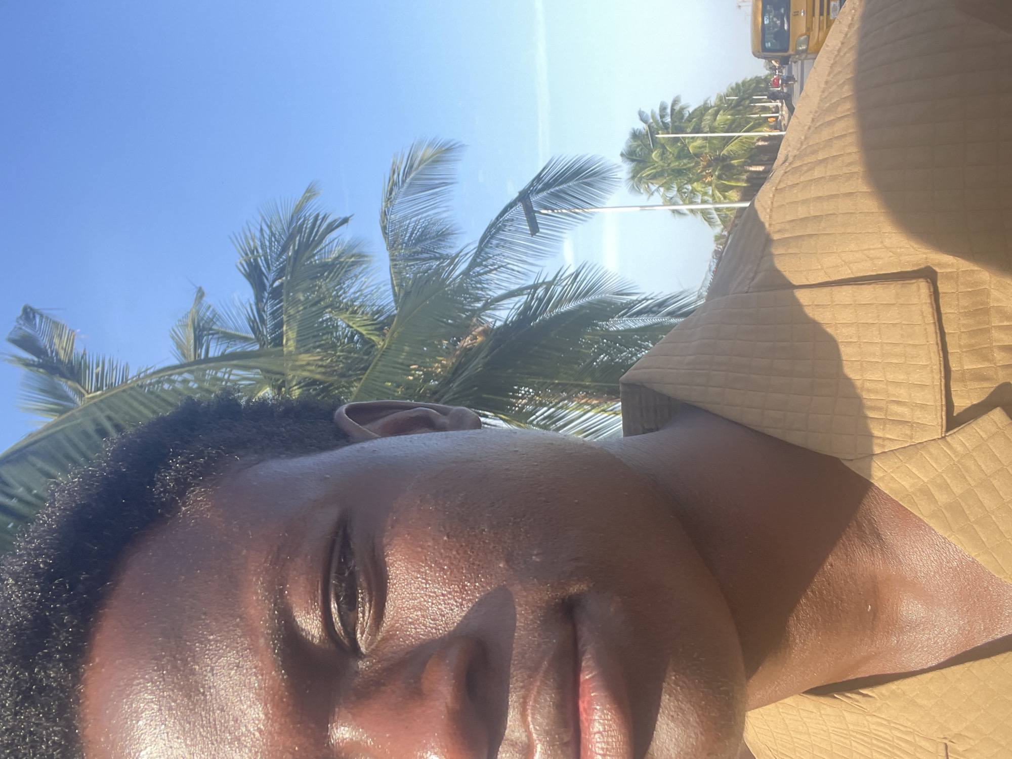 A close-up, rotated image shows a person's face with closed eyes. The face is sweaty and glistens in the sunlight. Behind the person are palm trees against a clear blue sky. In the upper right corner, a portion of a yellow bus and some buildings can be seen, suggesting a tropical urban environment. The presence of palm trees and the yellow bus indicates this might be Badagry, Nigeria, which is known for its beaches and tropical vegetation, and where yellow buses are common. The person is wearing a quilted-patterned tan colored garment.