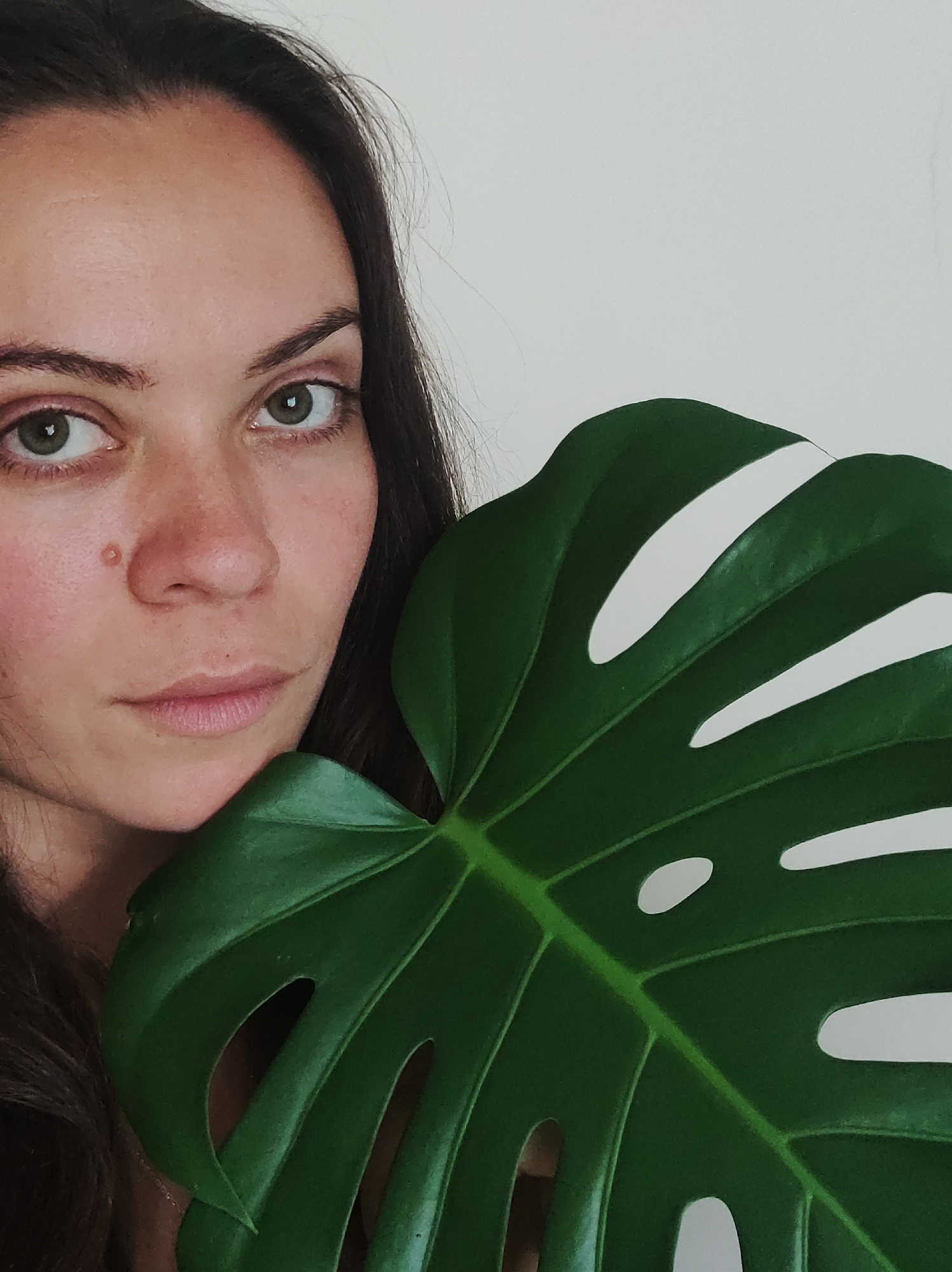 A close-up, waist-up shot features a woman holding a large, dark green Monstera leaf in front of her face, obscuring the right side of her head. She has dark brown hair, fair skin, and light green eyes. Her left eye is looking directly at the camera, while her right eye is partially visible. A small brown mole is present on her left cheek. The background is a plain, light-colored wall. The image does not provide any information about the city of Фоки, Russia.