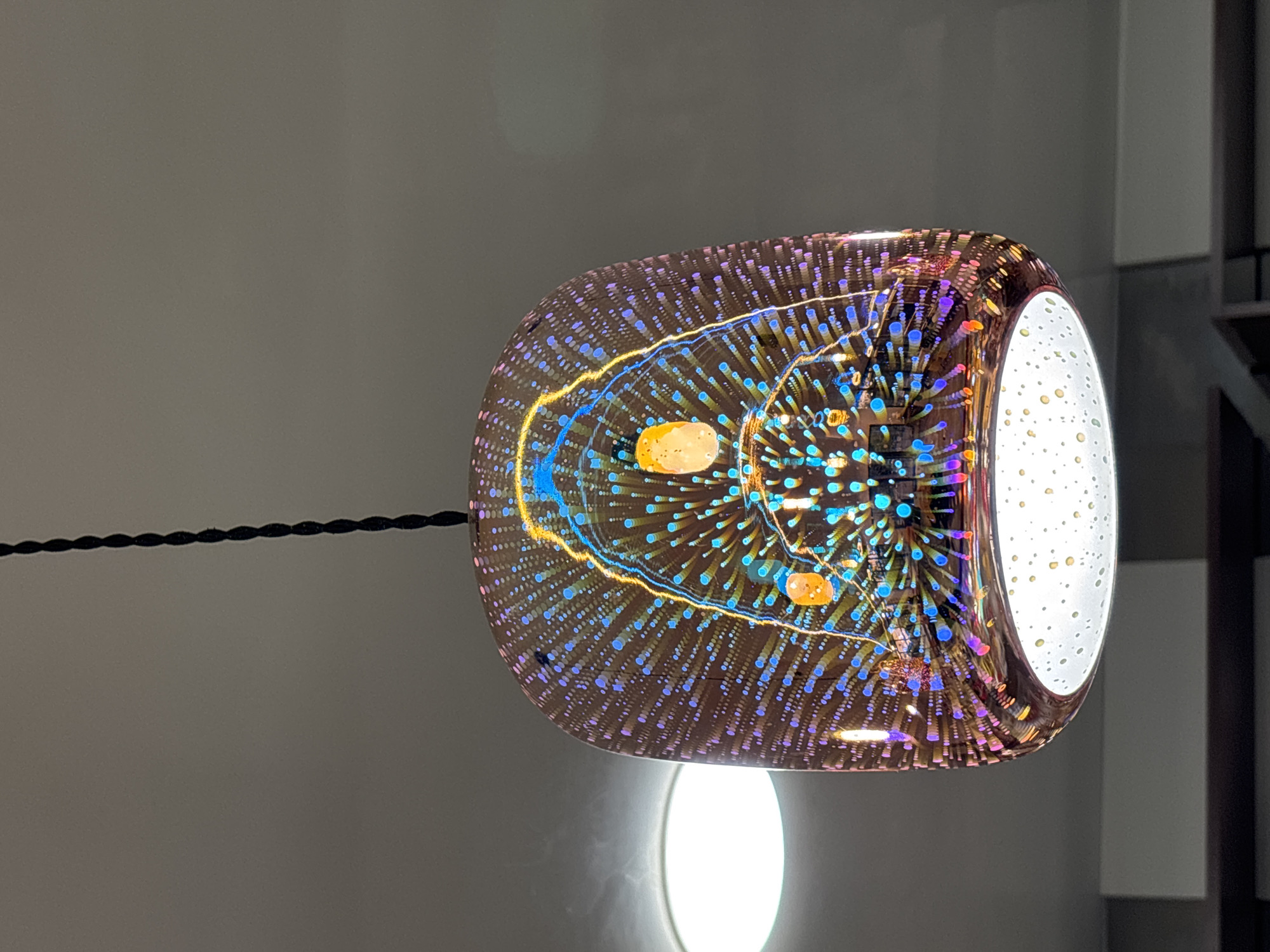 A decorative, cylindrical lamp with a complex internal pattern hangs from a braided cord. The lamp’s design features a dense grid of small, colorful dots and radiating lines in shades of blue, purple, and orange, creating a dynamic visual effect. An orange-colored shape can be seen inside. The lamp's right edge is a brighter white, dotted with pale yellowish spots. The lamp is positioned against a background of muted gray and white walls. Below the lamp is a circular, bright light fixture. The scene is inside a building. There is no information available in the image to determine its location in Gorna Malina, Bulgaria.