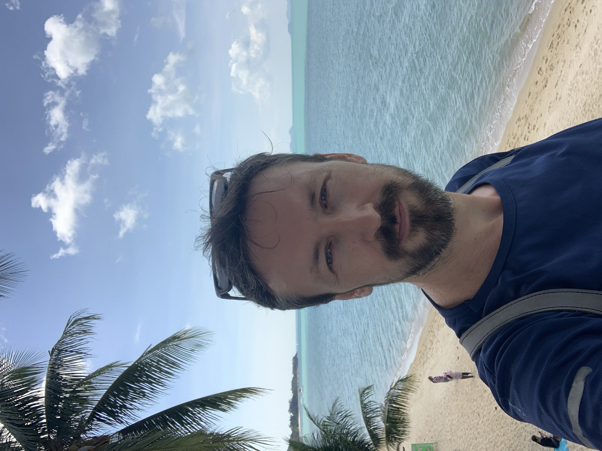 A man is depicted in a selfie, taken at a beach in Baan Mae Nam, Thailand. The image is oriented with a 90-degree clockwise rotation. The man, with dark hair and a beard, looks directly at the camera with a neutral expression. He is wearing a dark blue athletic-style top, and a pair of sunglasses is pushed up onto his head. A grey strap from a bag is visible over his right shoulder.
The background reveals a vibrant tropical scene. A clear blue sky with scattered white cumulus clouds dominates the upper portion. Below the sky, the calm, light turquoise water of the ocean stretches out, meeting a pale sandy beach where gentle waves break. In the distance across the water, a low-lying landmass or island can be seen. Several green palm trees are visible, with their fronds appearing in the lower left and bottom portions of the frame, suggesting the photo was taken from among them. Two small, indistinct figures are visible on the sandy beach further in the background, one standing and one closer to the water.
The time of day appears to be sunny daytime, indicating good weather for a beach outing. No legible text is visible in the image.