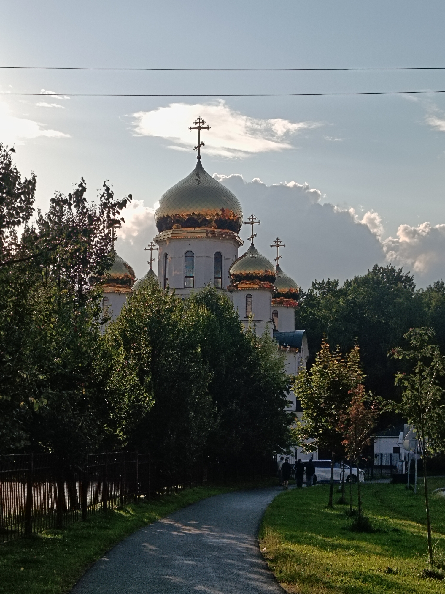 The image shows an Orthodox church with golden domes against a cloudy sky. In the foreground, there is a paved path curving to the right, bordered by a metal fence and green grass. Several trees with lush green foliage are scattered throughout the scene, some partially obscuring the church. In the background, a white car is parked, and three people are walking away from the viewer. The lighting suggests it is late afternoon or early evening, with sunlight casting shadows. The overall impression is one of a serene, possibly suburban, setting within Saint Petersburg, Russia.