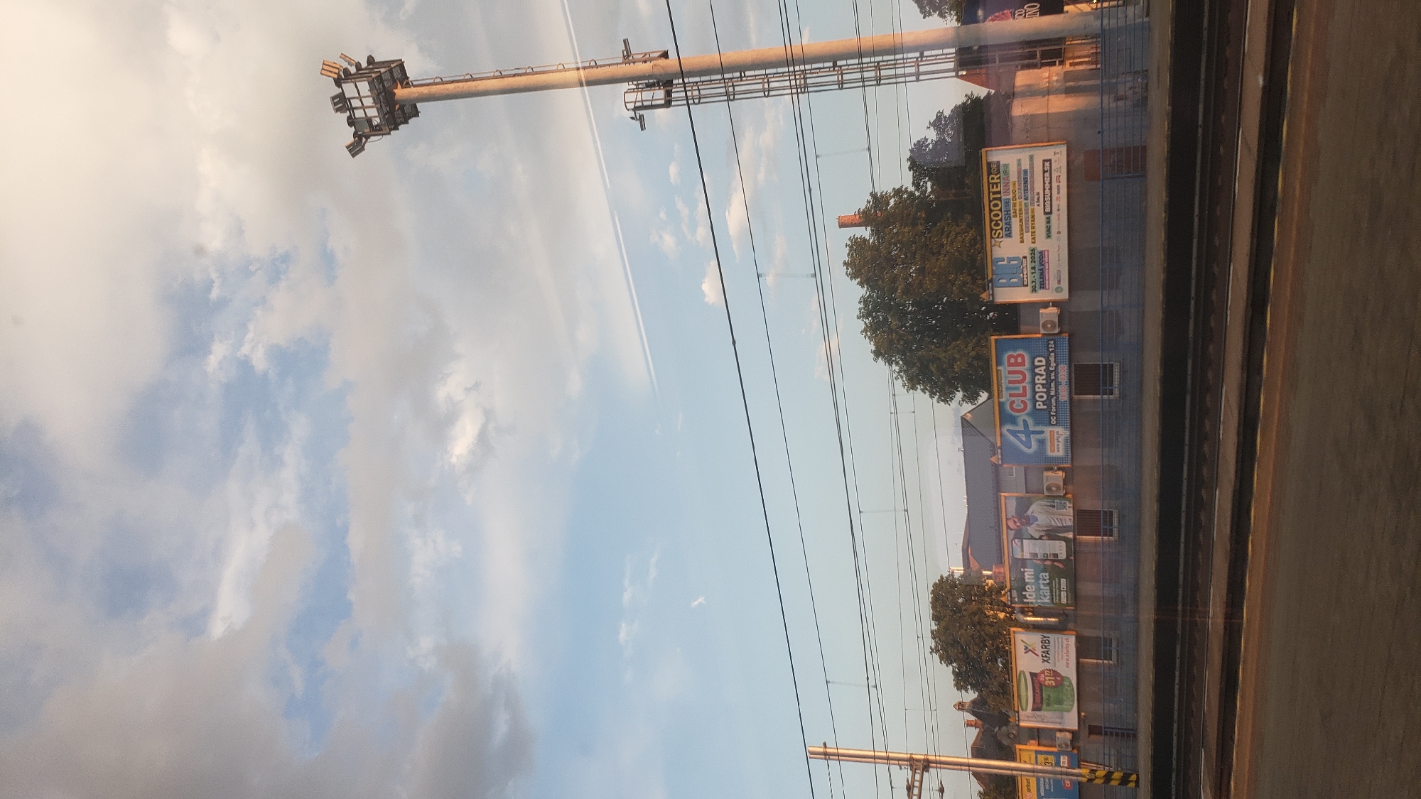 The image shows a street with a train track, billboards, and a lamppost. The sky is blue with clouds. On the billboards, there are advertisements for "XFARBY", "ide mi karta", "4 CLUB POPRAD", and "SCOOTER". There are also two buildings visible behind the billboards. One of the buildings has a red brick chimney. The train track is in the foreground of the image. The lamppost is tall and has lights on top. The scene appears to be in Poprad, Slovakia, as indicated by the "4 CLUB POPRAD" billboard.