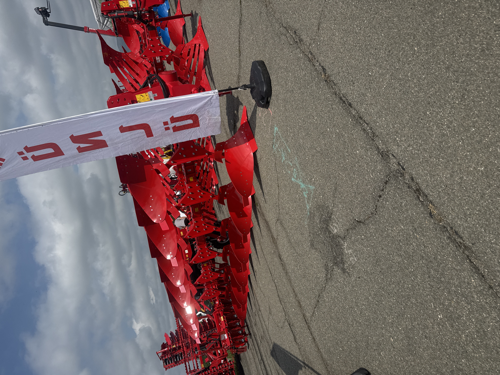 A large, bright red agricultural implement, resembling a multi-furrow reversible plow or cultivator, is displayed outdoors on a dark gray asphalt surface. The machinery consists of numerous red blades and structural components, with some distinct blue parts interspersed among the red elements, suggesting new equipment. A white flag bearing a red logo and partially visible text, appearing to be "U: L" (potentially reversed due to the flag's orientation), stands upright, attached to the machinery via a pole with a black base. The word "UMASAK" is clearly visible in white letters on a black arm extending from the top of the machinery.
The scene depicts an outdoor display, possibly at an agricultural exhibition or a dealership, in Stara Zagora, Bulgaria. The ground is paved asphalt with visible cracks and some light, ephemeral chalk marks. The sky is partially blue with significant white and gray cloud cover, indicating a daytime setting with overcast conditions. Shadows cast on the asphalt suggest moderate sunlight. A partial view of a person wearing a white shirt is discernible behind the machinery, hinting at human activity or presence around the display. The overall impression is one of a professional presentation of heavy-duty farm equipment.