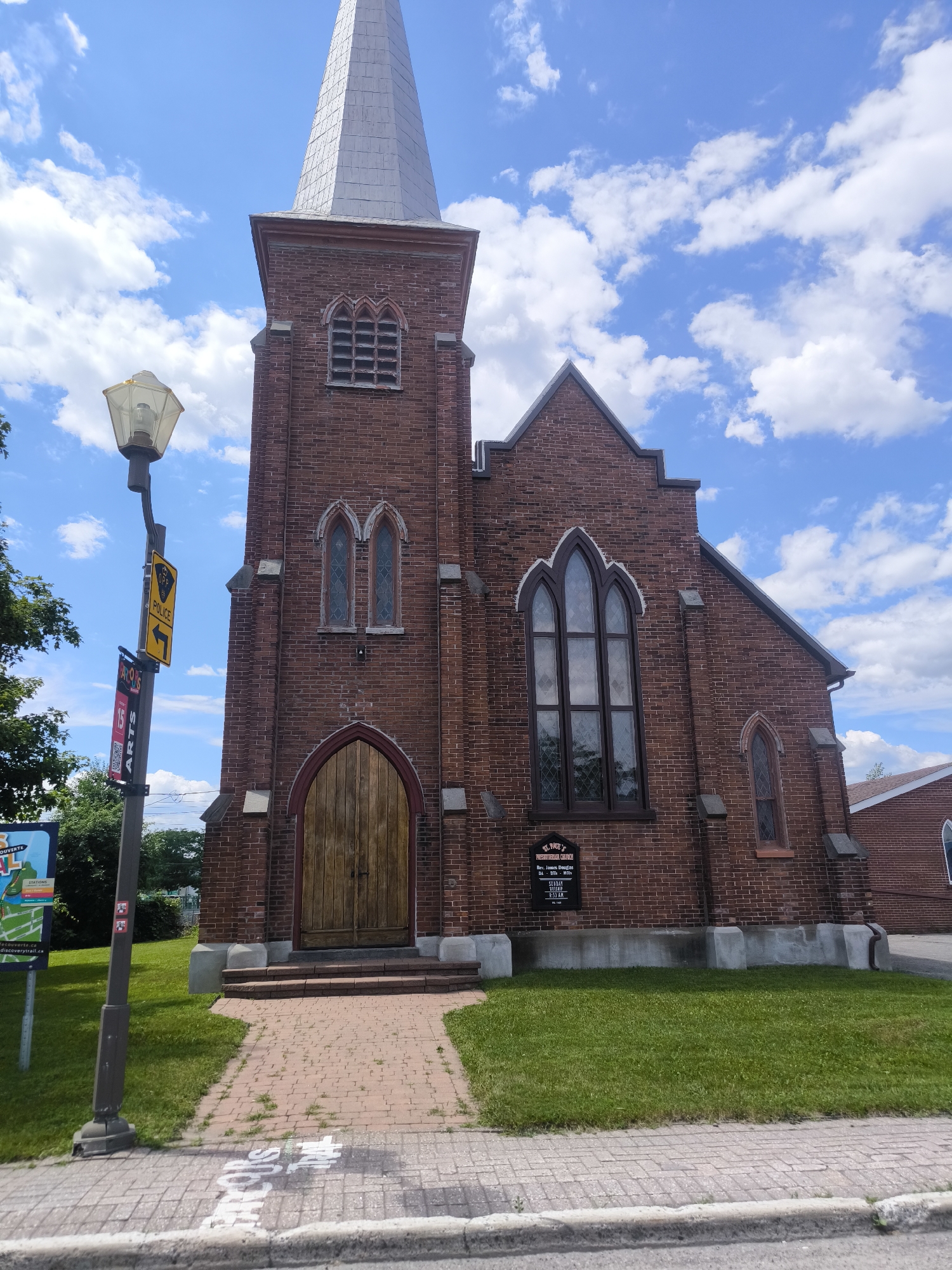 The image shows a red brick church with a tall, grey, pointed steeple in Hawkesbury, Canada. The church features gothic-style arched windows and a large wooden double door. A sign in front of the church reads "ST. PAUL'S PRESBYTERIAN CHURCH" and lists service times. A lamppost with several signs, including a yellow police sign, stands to the left of the church entrance. The church is surrounded by a green lawn and a paved walkway leading to the entrance. The sky is blue with scattered white clouds.