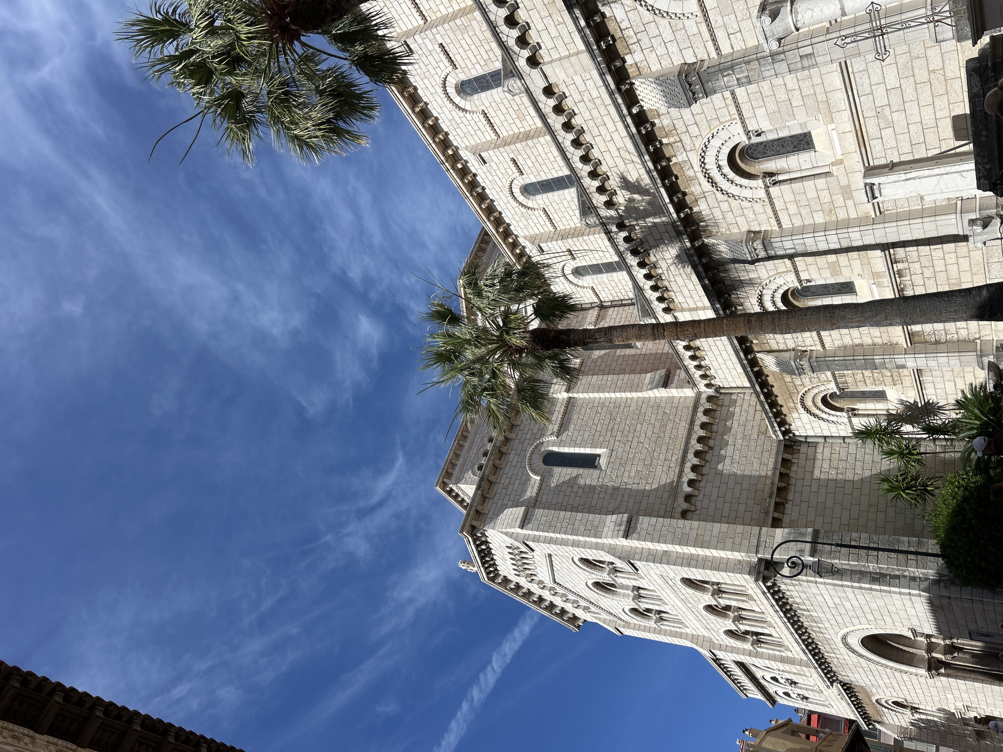 The image shows a low-angle view of the exterior of the Monaco Cathedral, also known as St. Nicholas Cathedral, located in Monaco-Ville, Monaco. The cathedral is constructed from light-colored stone blocks. It features multiple arched windows, some of which are stained glass. The architecture includes decorative cornices and buttresses. Two palm trees are prominently visible in the foreground and mid-ground, with their fronds reaching towards the sky. The sky is a vibrant blue with wispy white clouds, and sunlight casts shadows from the architectural features and palm trees onto the building. A street lamp is visible on the left side of the building. In the lower right corner, a metal cross is partially visible, suggesting the presence of a cemetery or memorial area adjacent to the cathedral. A few individuals are subtly visible in the lower left and lower right portions of the image, appearing to be pedestrians. The overall scene depicts a sunny day in Monaco.