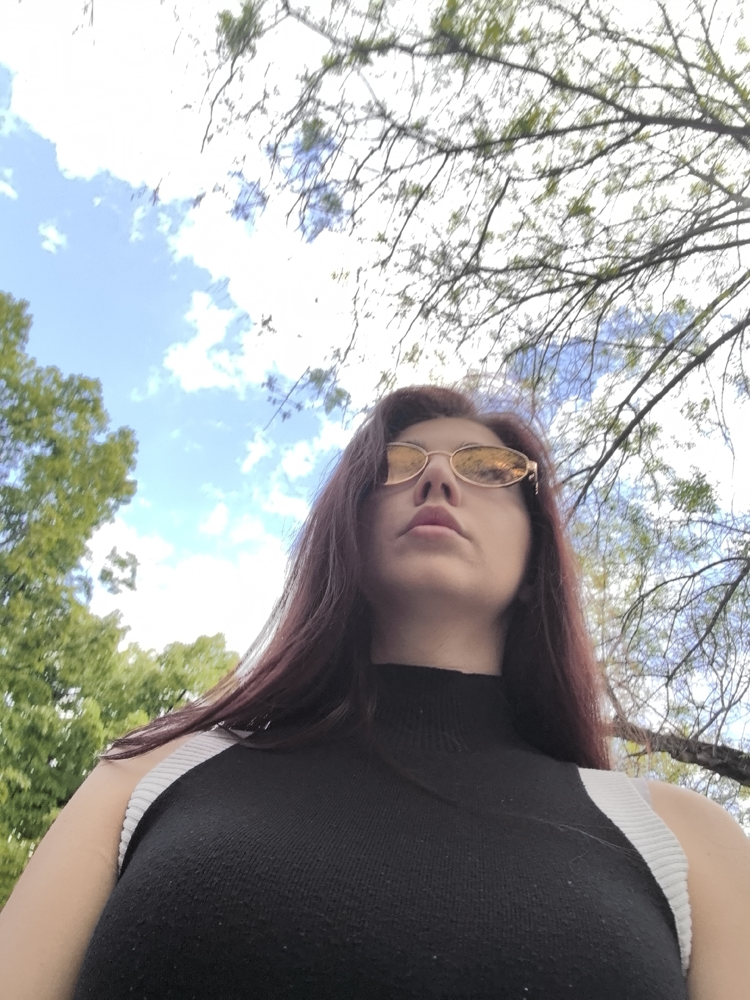 A low-angle, medium shot captures a woman in an outdoor setting. She wears a black, sleeveless top with white straps and gold-rimmed sunglasses. Her dark red hair frames her face, which is positioned slightly upwards. The background features trees with green leaves on the left, and a blue sky interspersed with white clouds above. Bare tree branches extend across the upper portion of the frame. The scene appears to be set in a park or garden. A definitive location in Stara Zagora cannot be determined from visual information alone.