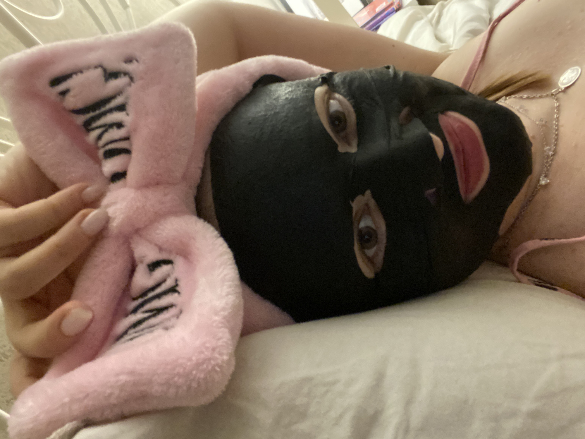 A person is reclining on a white pillow, wearing a black facial mask. The person has a pink fluffy hair wrap with black zebra stripes. The person is wearing a silver necklace and a pink top. The background is blurred and indistinct. There is no indication of Yekaterinburg, Russia, or any other specific location.
