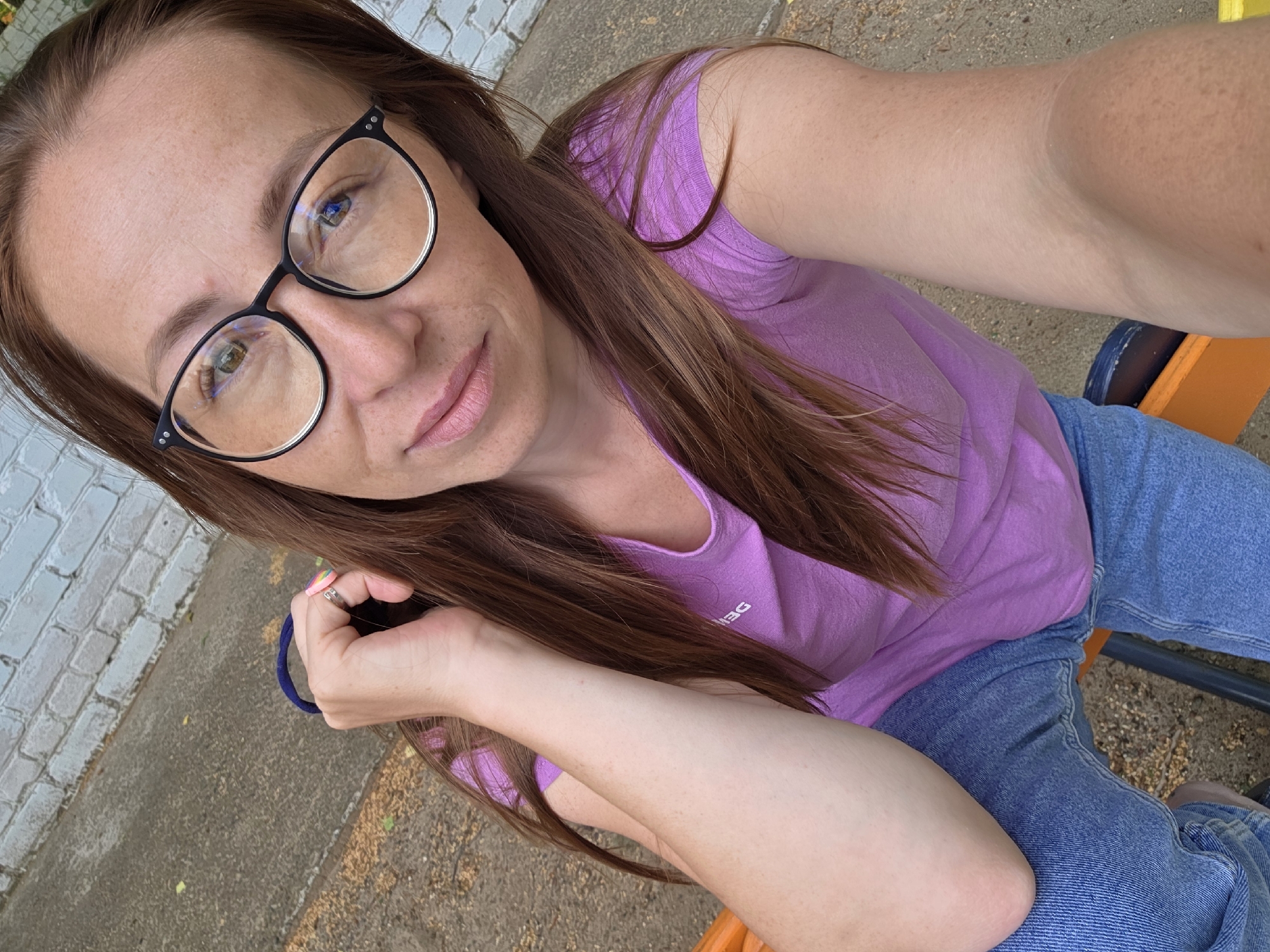 In Chaikovsky, Russia, a young woman with long brown hair and fair skin, wearing glasses, a pink t-shirt, and blue jeans, sits outdoors. She is holding a bag strap in her left hand and has a ring on her finger. The background consists of a grey concrete walkway and a white brick wall, indicating an urban setting. The lighting suggests it is daytime.
