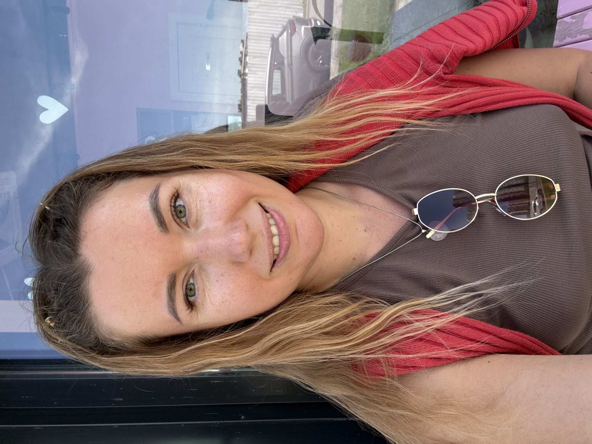 A woman is captured in a selfie, her face framed by blonde hair with blonde highlights. She wears a brown ribbed tank top with a red cardigan draped over her shoulders. A silver necklace with sunglasses hanging from it is visible. The background is a reflective surface, likely a window, revealing a blurred image of a car and a white fence. A white heart decal is also visible on the reflective surface. The overall lighting suggests an outdoor setting, possibly during the daytime.
