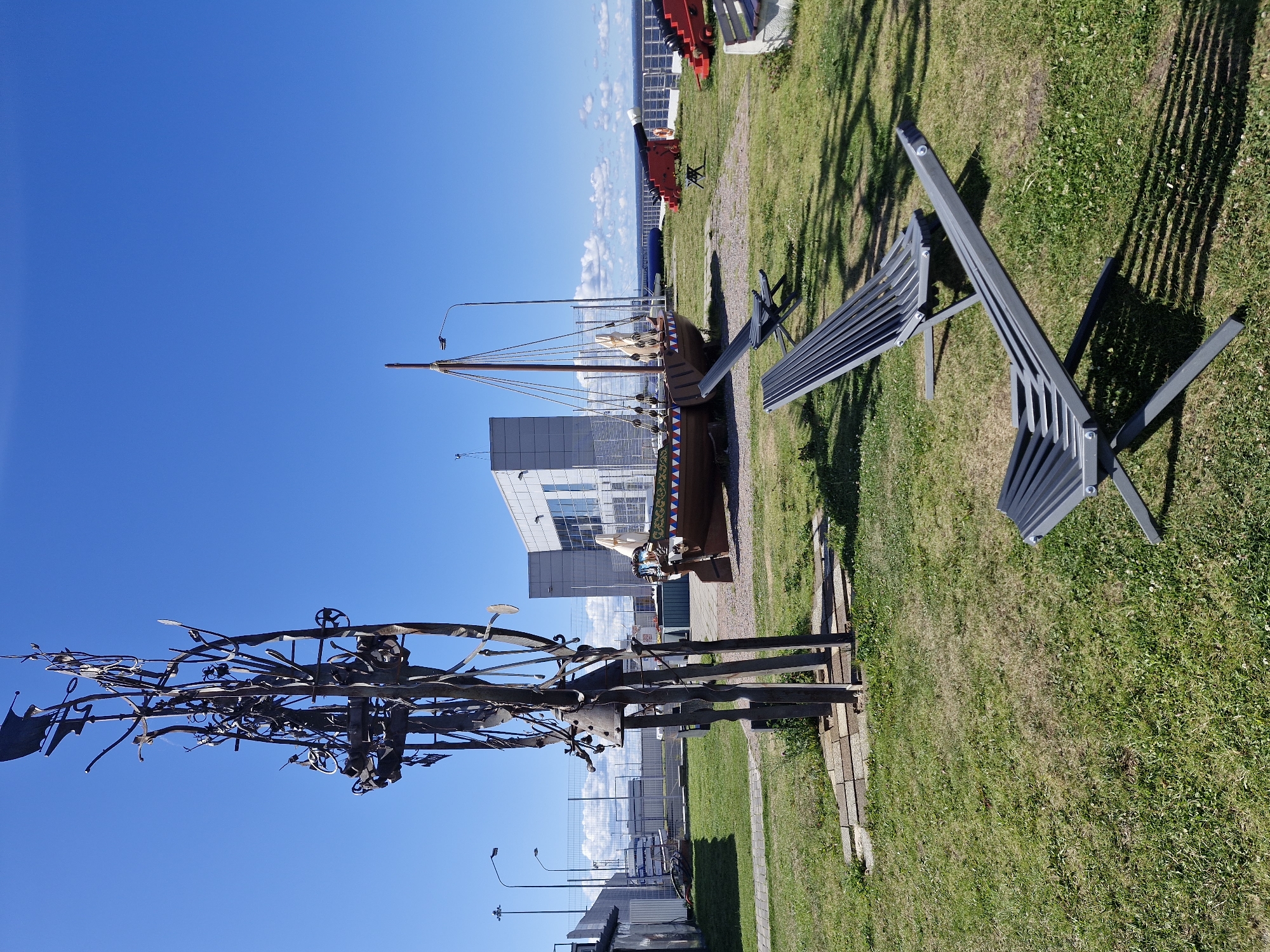 The image depicts an outdoor area on a clear, sunny day with a bright blue sky. In the foreground, two dark grey, slatted lounge chairs are positioned on a grassy lawn. Behind them, a large, intricate metal sculpture stands on a paved base. To the right of the sculpture is a wooden boat with a mast and sail, decorated with a triangular pattern along its hull. Further in the background, a modern building with a glass facade is visible. Several red cannons are also present on a paved area, some facing towards a body of water, likely the Gulf of Finland. The overall scene suggests a historical or maritime-themed exhibition or park. This location is the Rurik Monument and Museum, situated on the Petrograd Side of Saint Petersburg, Russia, overlooking the Gulf of Finland.