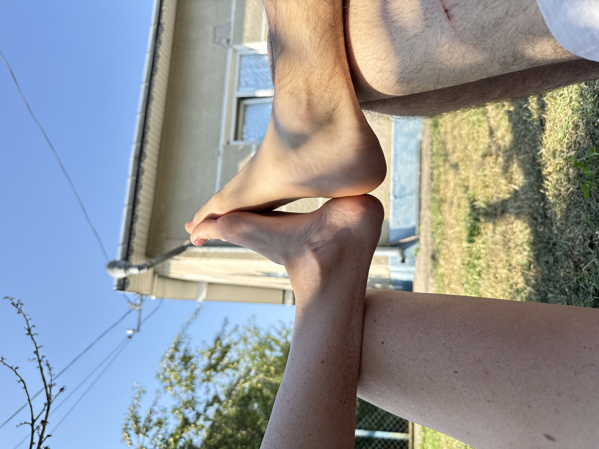 The image shows a close-up of two pairs of feet touching toes in front of a house. The feet belong to two individuals, one on the left and one on the right. The person on the left has fair skin with freckles. The person on the right has tanned skin and visible leg hair. The background shows a light-colored house with a grey roof and a window. There are trees and a blue sky above, suggesting an outdoor setting. The ground is covered in grass and some shadows are cast. The scene appears to be in a residential area, likely a backyard.