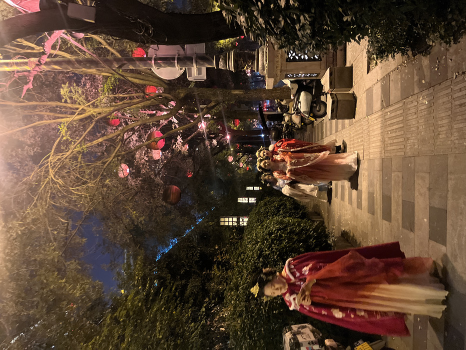 This nighttime image depicts a paved pedestrian walkway in Yanta District, China. Five individuals, identified as adult females, are walking from right to left along the path. All five are dressed in elaborate, layered traditional Chinese garments (Hanfu), featuring predominantly red, white, pink, and cream colors, with decorative headpieces. The foreground figure, positioned on the left, wears a red outer cloak over a lighter gown and is observed looking down at a white electronic device held in her hands. The four individuals behind her are in various traditional ensembles, with one central figure in pink and white looking forward.
The walkway is lined on both sides by dense green foliage and trees. Numerous spherical red and orange lanterns are suspended from the tree branches overhead, providing ambient illumination. A dark street lamp pole stands on the right side of the path, with a scooter or moped parked nearby. In the background, visible through the foliage on the left, is a building facade with multiple illuminated square window-like features. The path itself is constructed from rectangular paving stones, with some tactile strips discernible. The overall lighting is dim, sourced from the lanterns, streetlights, and background building lights.