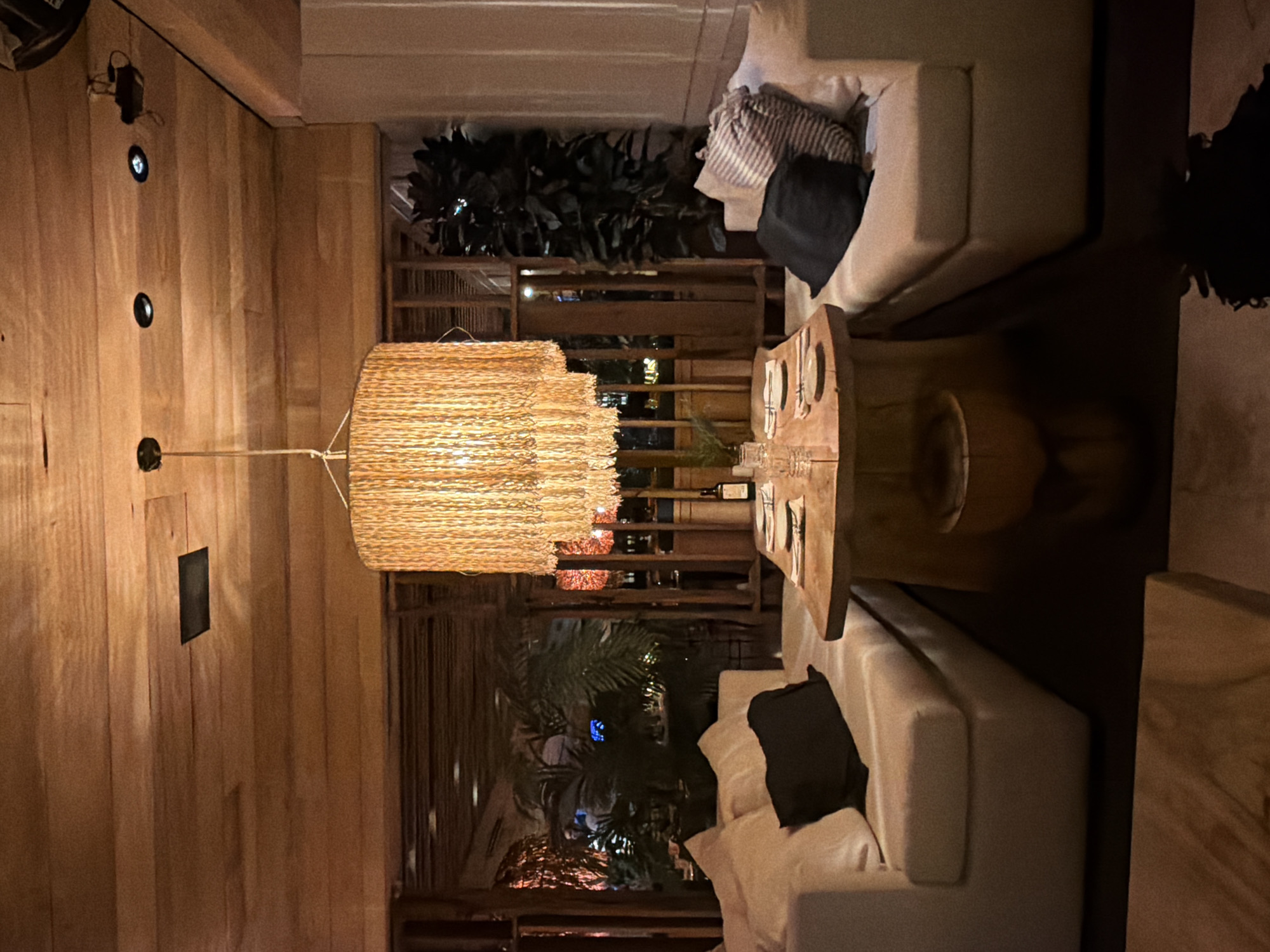 This image appears to be an interior shot of a dining area, likely in a restaurant. The most prominent feature is a large, tiered, woven straw or rattan chandelier hanging above a wooden dining table set with plates and glasses. There is a wine bottle on the table as well. Two light-colored sofas with pillows are positioned around the table, indicating a lounge or more relaxed dining setting. Behind the dining area, there is a view through a lattice or screen, showing greenery, suggesting an outdoor or semi-outdoor space. The walls of the interior appear to be paneled with wood. There are also small recessed lighting fixtures on the wall. Given the style of the decor and architecture, it is possible that this is a location in Buenos Aires, Argentina, as the city has numerous restaurants and bars with stylish and modern interiors, often incorporating natural materials and outdoor/indoor design elements.