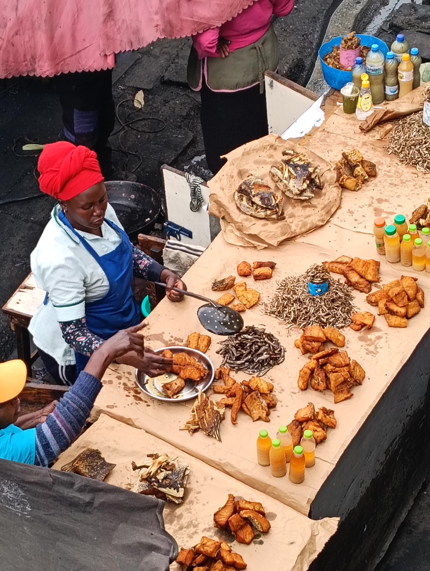 The image depicts a bustling outdoor market scene in Nairobi, Kenya, likely a fish market given the prominent display of various fish products. In the foreground, a woman wearing a red headscarf and a blue apron is actively engaged in handling a plate of fried fish. Her hands are visible as she interacts with another person, whose hands are also shown reaching for the fish.
The market stall is laden with a variety of goods, primarily food items. Piles of dried fish, some whole and others filleted, are spread across brown paper. There are also numerous pieces of fried fish, appearing golden brown and crispy. Several small bottles of liquids, possibly condiments or beverages, are arranged on the stall. In the background, other market-goers can be seen, with one individual wearing a pink garment and another in a dark blue shirt and black trousers. The ground appears to be a rough, dark surface, characteristic of an outdoor market. The overall impression is one of lively commerce and food preparation.