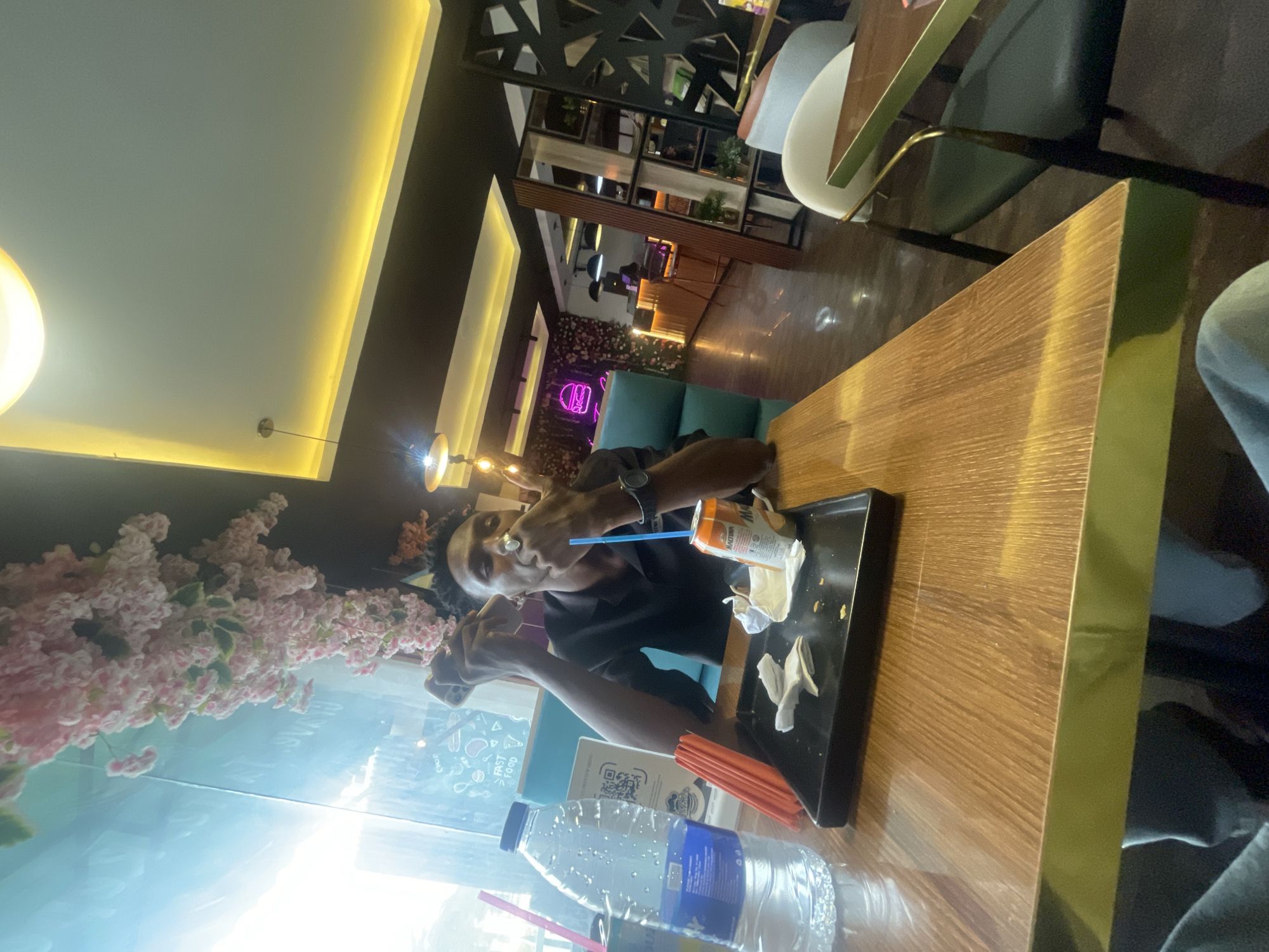 The image shows a person sitting at a table in a restaurant, presumably in Adankolo, Nigeria. The person is wearing a black shirt and has short hair. They are holding a blue straw and appear to be drinking from a beverage can on a black serving tray. The tray also holds paper napkins and what appears to be remnants of food. A bottle of water and additional straws are on the table. The table is made of wood and has a metallic gold border. The background features a decorative wall with flowers and a view outside of a street with buildings. Other tables and chairs are visible in the background, indicating a restaurant setting.