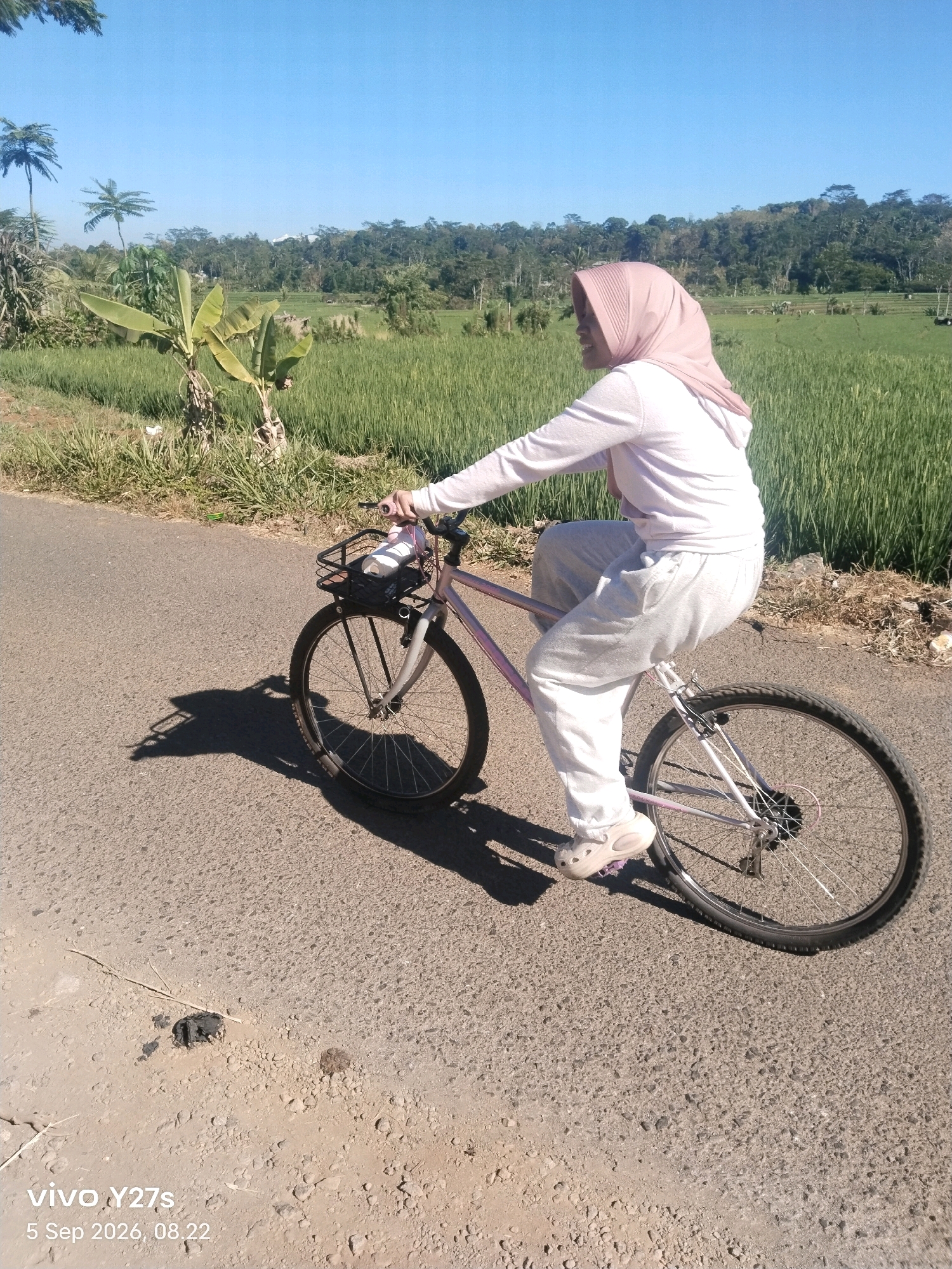 A young woman wearing a pink hijab, a white long-sleeved shirt, and white sweatpants rides a pink bicycle on a paved road. She is looking to her right, with a faint smile visible. The bicycle has a black basket on the front containing a white object. Her shadow is cast on the road to her left. The road is surrounded by green vegetation, including rice paddies and banana trees, with a line of trees and hills in the background. The sky is clear and blue. The timestamp in the lower left corner indicates "vivo Y27s 5 Sep 2026, 08.22", suggesting the photograph was taken in Selojetis, Indonesia, on September 5, 2026, at 8:22 AM.