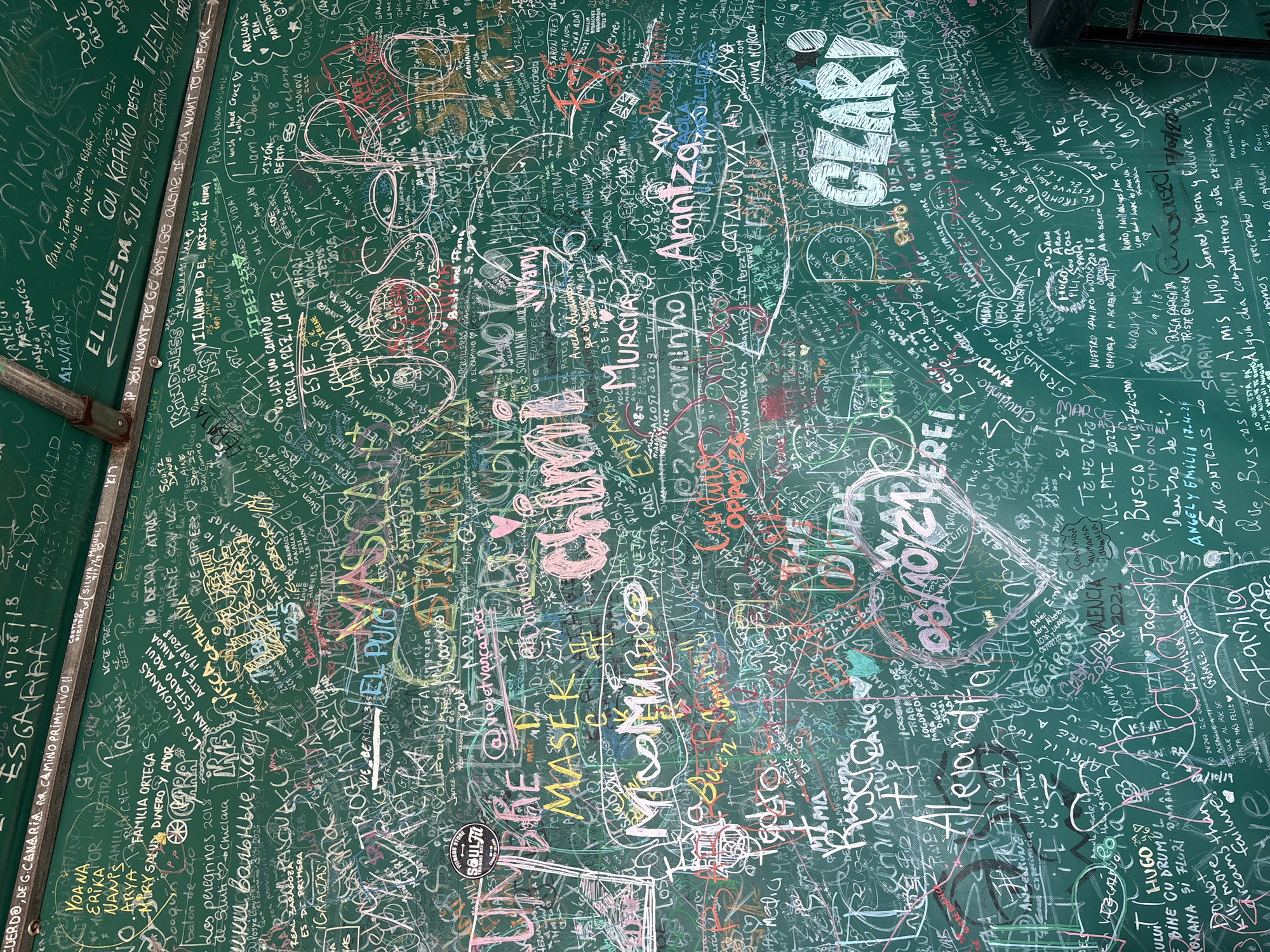 The image shows a densely covered wall in Lengüelle, Spain, entirely filled with graffiti. The wall appears to be part of a public space, potentially associated with the Camino de Santiago pilgrimage route. The graffiti consists of names, dates, messages, and drawings in various colors and styles, written in different languages. It appears to be a collection of expressions left by pilgrims passing through the area. The surface is a deep green color with the graffiti covering every inch of available space. The presence of visible writing in a diverse range of alphabets and languages strongly suggests an international array of visitors.