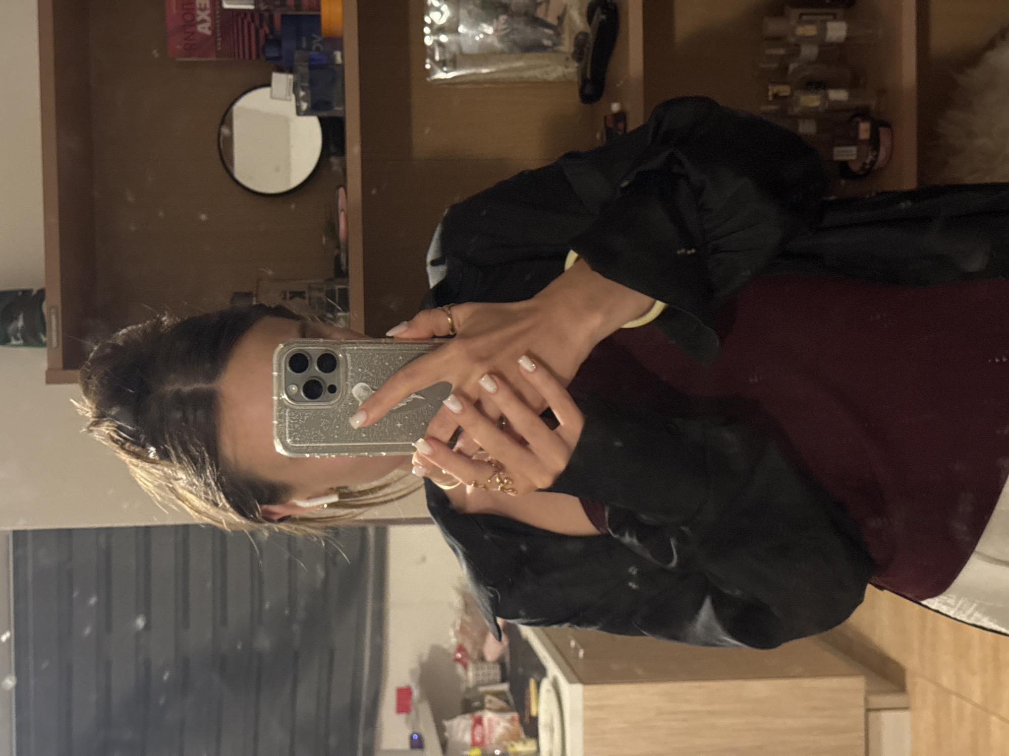 A young woman with her hair pulled up in a bun takes a selfie in a mirror, wearing a dark jacket over a burgundy top and white pants. She has white nail polish and is wearing earbuds and rings on her fingers. The background shows a shelf with various items, including bottles of perfume and a small mirror. To the left, a kitchen counter is visible with some items on it. The scene appears to be indoors, likely in a residential setting. There is no information to suggest this location is in Budva, Montenegro.