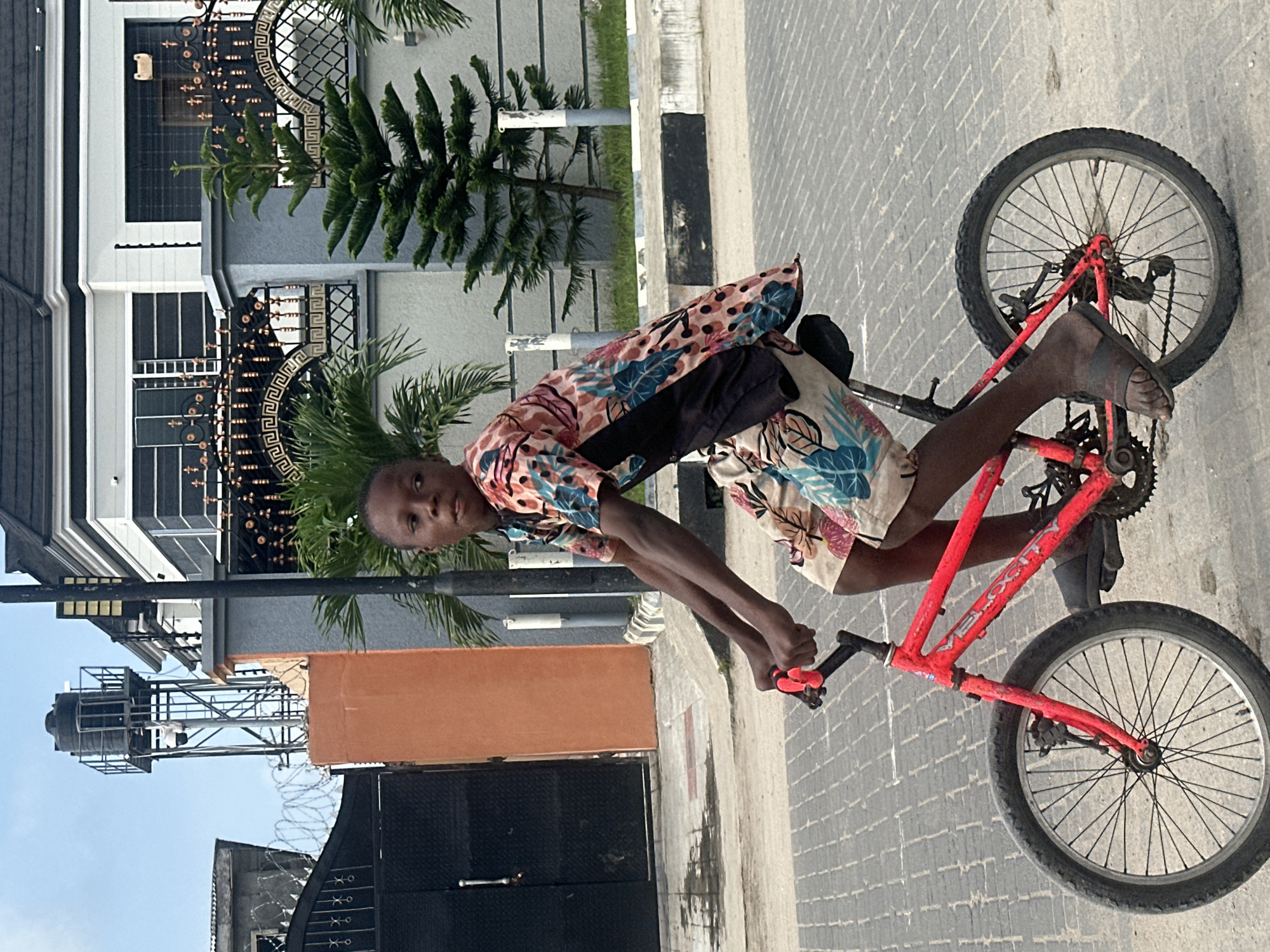 In Badagry, Nigeria, a young Black boy is captured mid-action, riding a small, bright red bicycle on a paved street. He is wearing a colorful short-sleeved shirt with a tropical leaf pattern and matching shorts. The bicycle has black tires and spokes, a black seat, and red handlebars with black grips. He is wearing dark-colored sandals.
The background features a modern, multi-story building with a grey facade, white trim, and decorative metal fencing. To the left, there is a tall, orange-colored structure topped with a black water tank and surveillance cameras. A large black metal gate with elaborate scrollwork is visible. Palm trees and a coniferous tree are planted in front of the building, adding a touch of greenery to the urban landscape. The sky is a pale blue, indicating daytime. The street is paved with interlocking brick pavers.