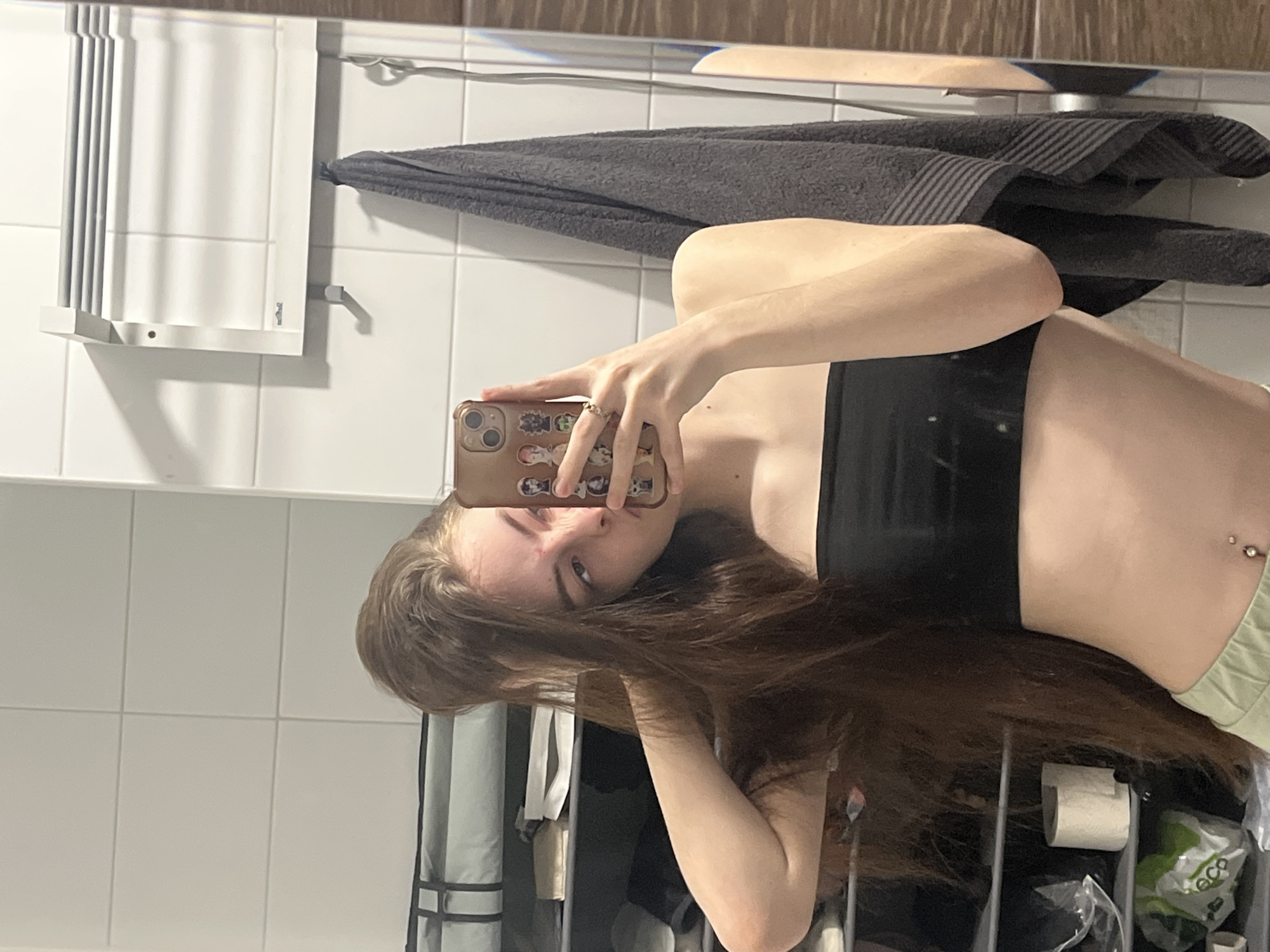 The image is a mirror selfie taken in what appears to be a bathroom, with the photo oriented 90 degrees clockwise. The subject is a young individual, likely female, with long, dark brown hair partially obscuring her face and arm. She is wearing a black crop top and light green bottoms, and has a navel piercing visible on her abdomen. She holds a smartphone, encased in a light brown/gold case decorated with small cartoon stickers, to capture her reflection.
The background, visible in the mirror, shows white tiled walls. Mounted on the wall is a white, slatted object, possibly a towel warmer or shelf. A dark grey/charcoal colored towel is draped over a rail above this object. Another rail is visible further up. In the bottom right corner of the reflection, a roll of toilet paper is seen alongside some indistinct items, including plastic packaging with the text "eco" visible. The overall setting is functional and domestic, consistent with a bathroom environment. The individual's expression is neutral as she looks into the phone. The photograph is set in Saint Petersburg, Russia.