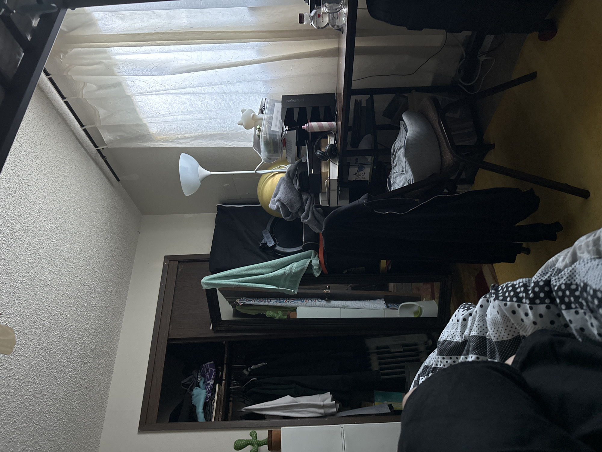 The image depicts a cluttered bedroom. On the left, a closet is partially open, revealing clothes hanging on a rack and items stacked on shelves. A small potted cactus is visible on top of a white dresser next to the closet. In the center of the room, a desk is covered with various items including papers, a lamp, headphones, and bottles. A chair sits in front of the desk, with a gray cushion on it. A large window with sheer curtains is on the right side of the room. The floor is covered with a yellow carpet. A person is partially visible in the foreground, lying on a bed with a patterned blanket. The overall impression is one of disarray and a lived-in space. There is no information in the image to suggest the location is in Vancouver, United States of America.