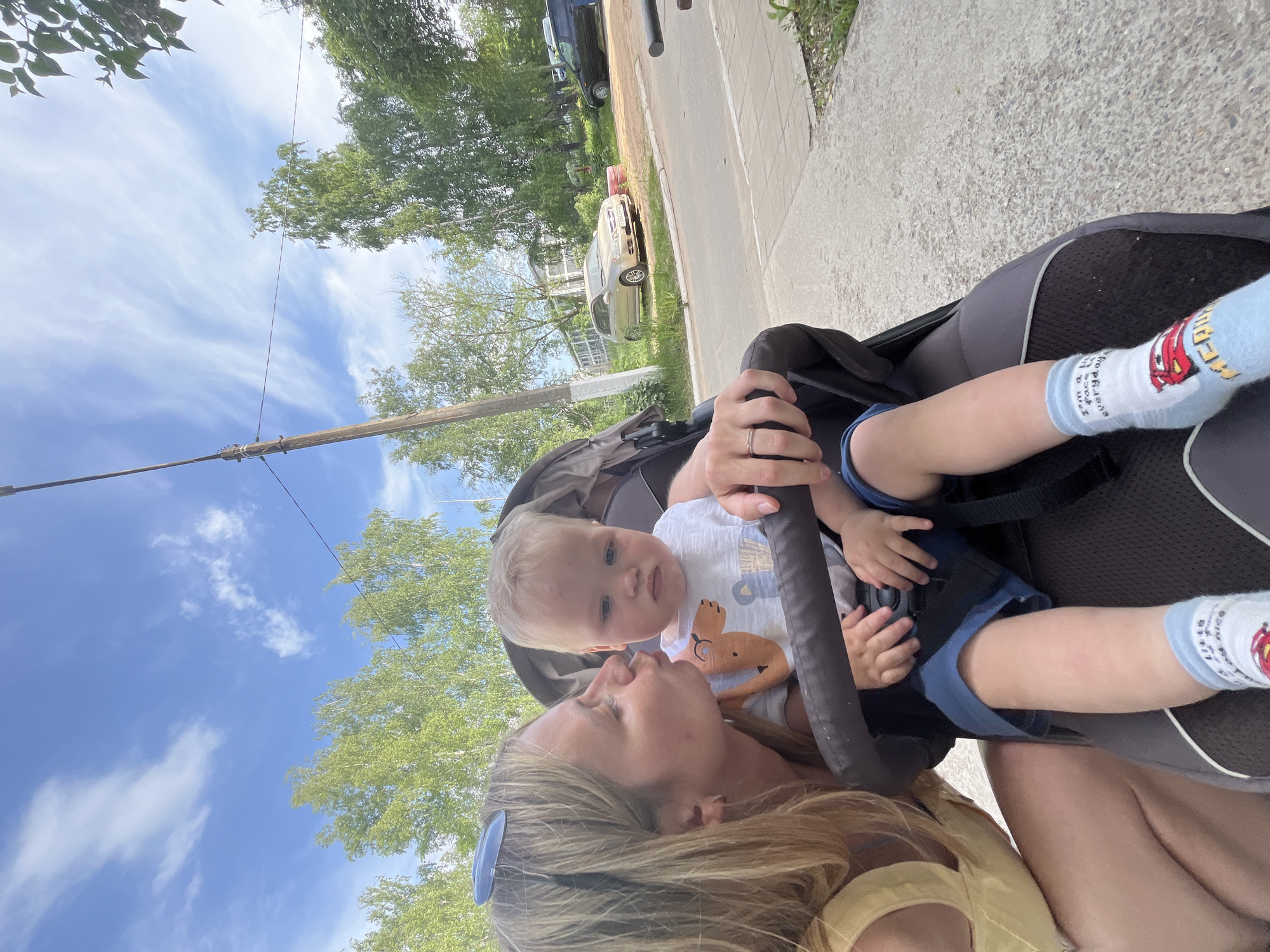 A medium, eye-level shot shows a woman and a baby in a stroller on what appears to be a sidewalk in Chaikovsky, Russia. The woman, with blonde hair and wearing a yellow top, is looking at the baby. The baby, with blonde hair, is wearing a white t-shirt with a cartoon character and blue socks with a car image on them. The baby is strapped into the stroller. In the background, there is a road with a silver car and a dark-colored SUV parked on the side. Trees and a clear blue sky with scattered clouds are also visible. A utility pole is seen on the left side of the image.