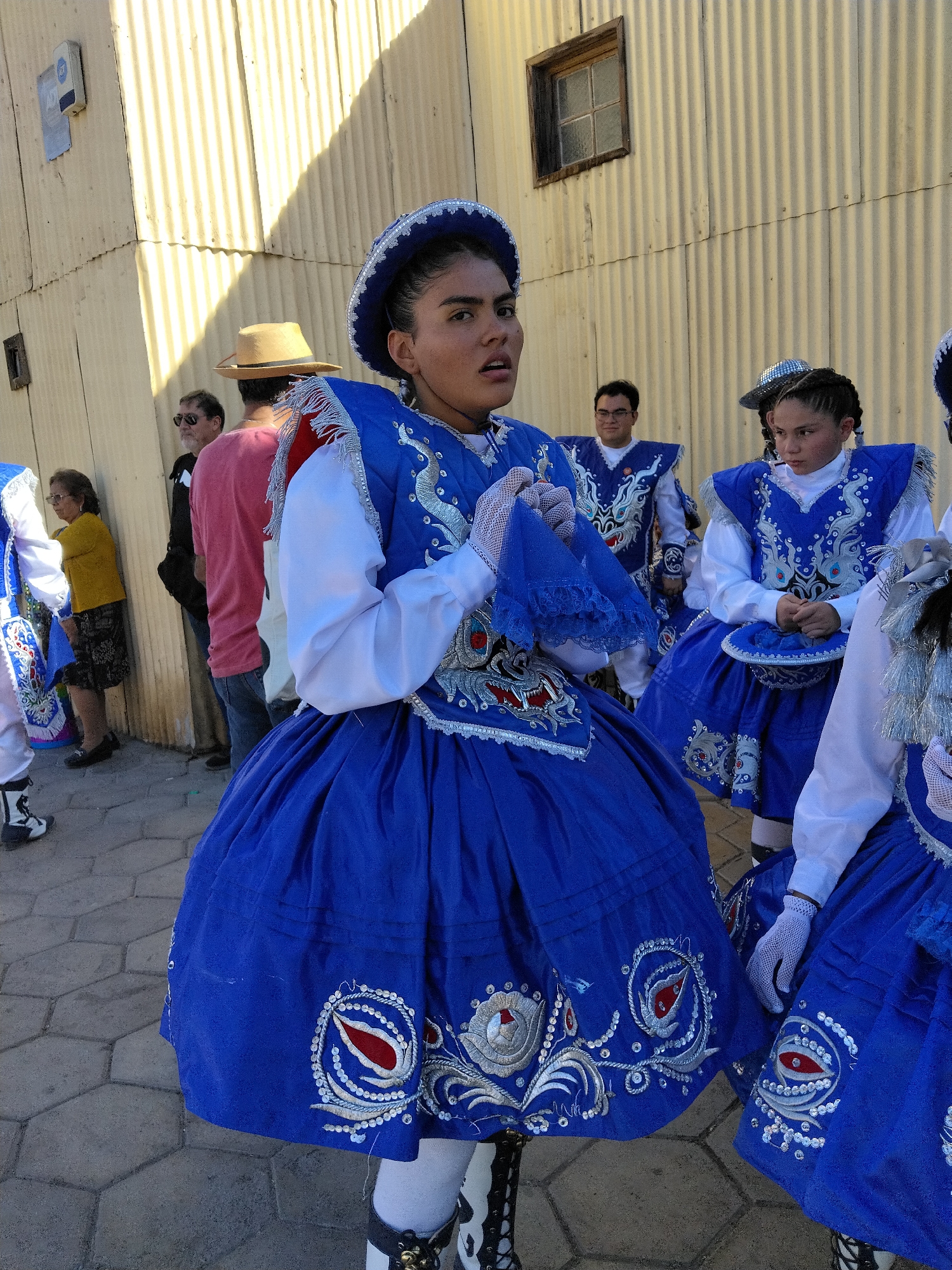 The image depicts a street scene in Pozo Almonte, Chile, featuring a group of people, predominantly young women, dressed in elaborate traditional blue and white costumes. The costumes include full skirts, white blouses with intricate silver and red embroidery, and decorative headwear. One young woman in the foreground is wearing a blue hat with white trim, a blue skirt with elaborate white and red embroidered circular motifs, and white gloves. Her expression is directed towards the camera with her mouth slightly open. Behind her, other individuals in similar attire are visible, including a person wearing a blue and white patterned hat. In the background, there are several adults dressed in casual clothing, including a man in a tan hat and a red shirt, and another man in sunglasses and a black shirt. The setting appears to be an outdoor area with a textured gray pavement and a corrugated metal wall in the background, which has a small, square window and a shadow cast upon it. The overall impression is of a cultural event or performance taking place in the city.