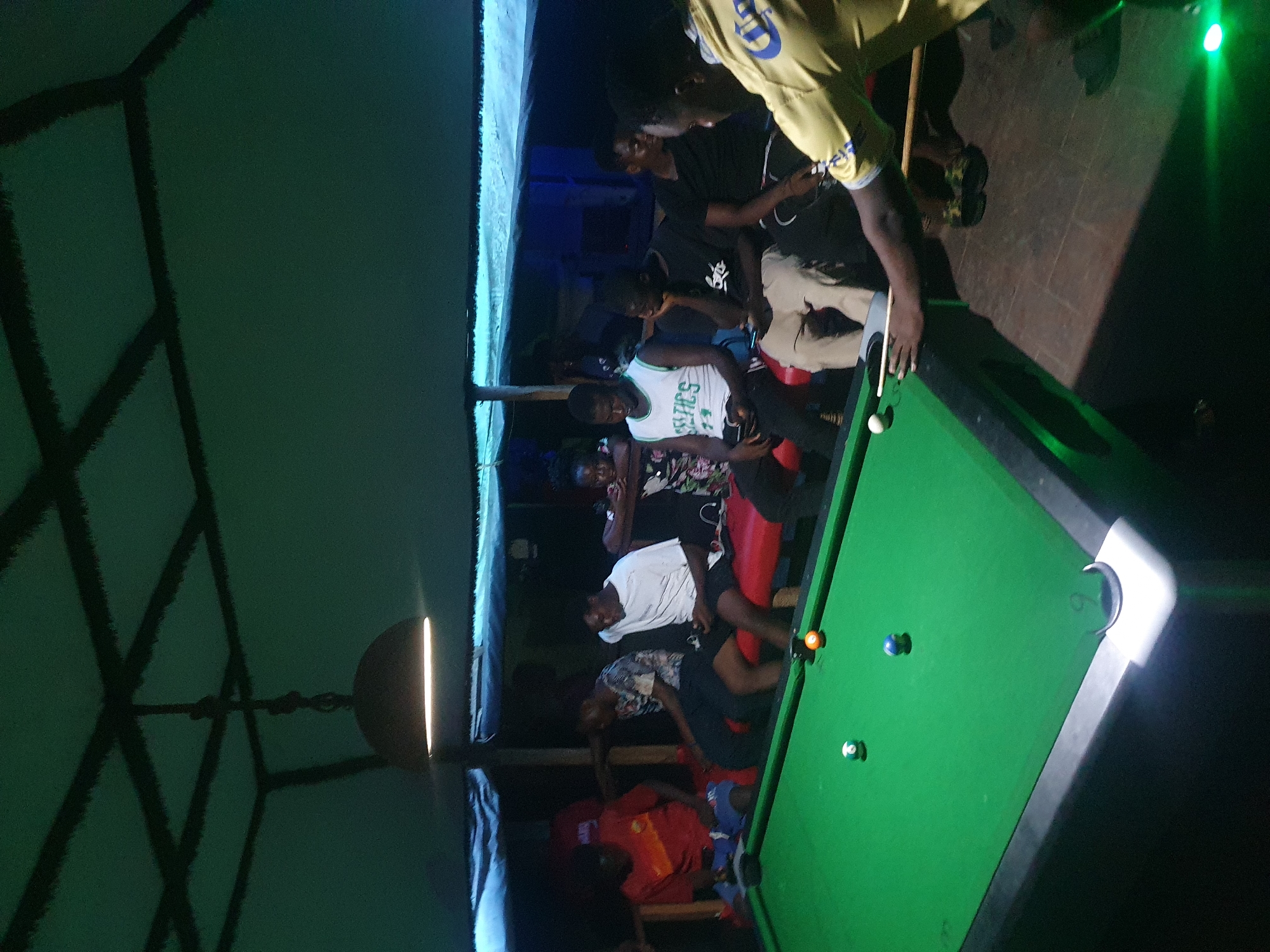 The image captures an indoor scene, likely in the evening or at night, where a group of young adults is gathered around a pool table. The setting appears to be a dimly lit recreational area, possibly a bar or game room, with ambient green lighting and sparse direct light sources. The image is oriented vertically, with the main activity occurring on the right side of the frame.
In the foreground, a person wearing a yellow and blue shirt is actively playing pool, leaning over the green felt table with a cue stick in hand. A white cue ball and several colored balls (orange/yellow and blue) are visible on the table. Other individuals are either sitting on benches or standing nearby, observing the game. One person seated on a red bench in the mid-ground is wearing a white tank top with "CELTICS" and a number, possibly "74," printed on it. Other spectators wear casual attire, including t-shirts and shorts.
The atmosphere seems relaxed and social, focused on the game of pool. The floor beneath the players appears to be tiled or brick-paved. On the far left, a wall features dark, geometric patterns against a dark green background. A light fixture with a bright, warm glow is visible on this wall. Another bright light illuminates one of the pool table's corner pockets. The overall impression is one of leisure and camaraderie within a local establishment in Lokoja, Nigeria.