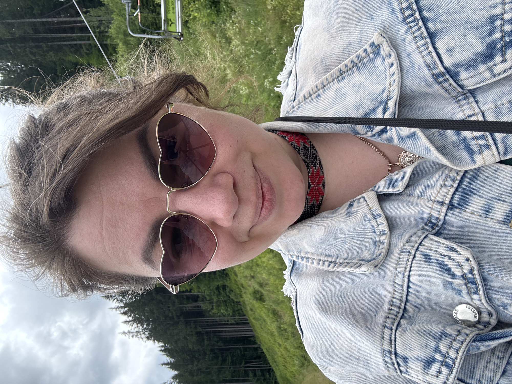 This is a selfie of a woman outdoors. She is wearing a light-wash denim jacket and heart-shaped sunglasses. A beaded choker necklace with red and black patterns is visible around her neck, along with a silver necklace with a pendant. Her hair is styled in loose waves and appears to be pulled back.
The background shows a lush green hillside with trees, suggesting a natural, possibly mountainous, environment. A ski lift structure is visible in the upper left corner, along with a cable stretching across the frame. The sky is cloudy. The scene appears to be set during the daytime, with overcast weather.
A button on the woman's denim jacket has the text "VIVIENNE WESTWOOD LONDON" inscribed on it. The overall impression is one of casual leisure in a scenic outdoor location.