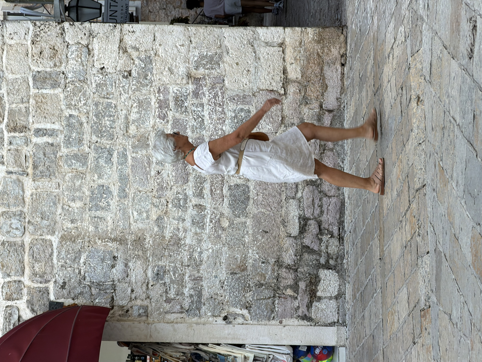 A woman with short, gray hair is captured mid-stride on a cobblestone path. She is wearing a simple, white, knee-length dress, a beige belt cinched at her waist, and tan flat sandals. A turquoise beaded necklace adorns her neck. Her left arm is extended outwards and slightly upwards, while her right arm is bent at the elbow, her hand near her waist. Her body is angled, suggesting movement, and her gaze is directed forward and slightly down.
The background features a rough, weathered stone wall constructed from irregularly shaped blocks of varying gray and white tones. To the left, partially visible, is a dark red awning and a display of what appears to be books or magazines. Above the stone wall, a sign with white text on a dark background is visible; the word "UNTA" is clearly legible, with "ONOSBA" and "STAURANT" also discernible below it, hinting at a restaurant or establishment. The lighting suggests it is daytime, with soft shadows. The scene appears to be outdoors in an old town setting, possibly in Budva, Montenegro, given the stone architecture and the general ambiance.