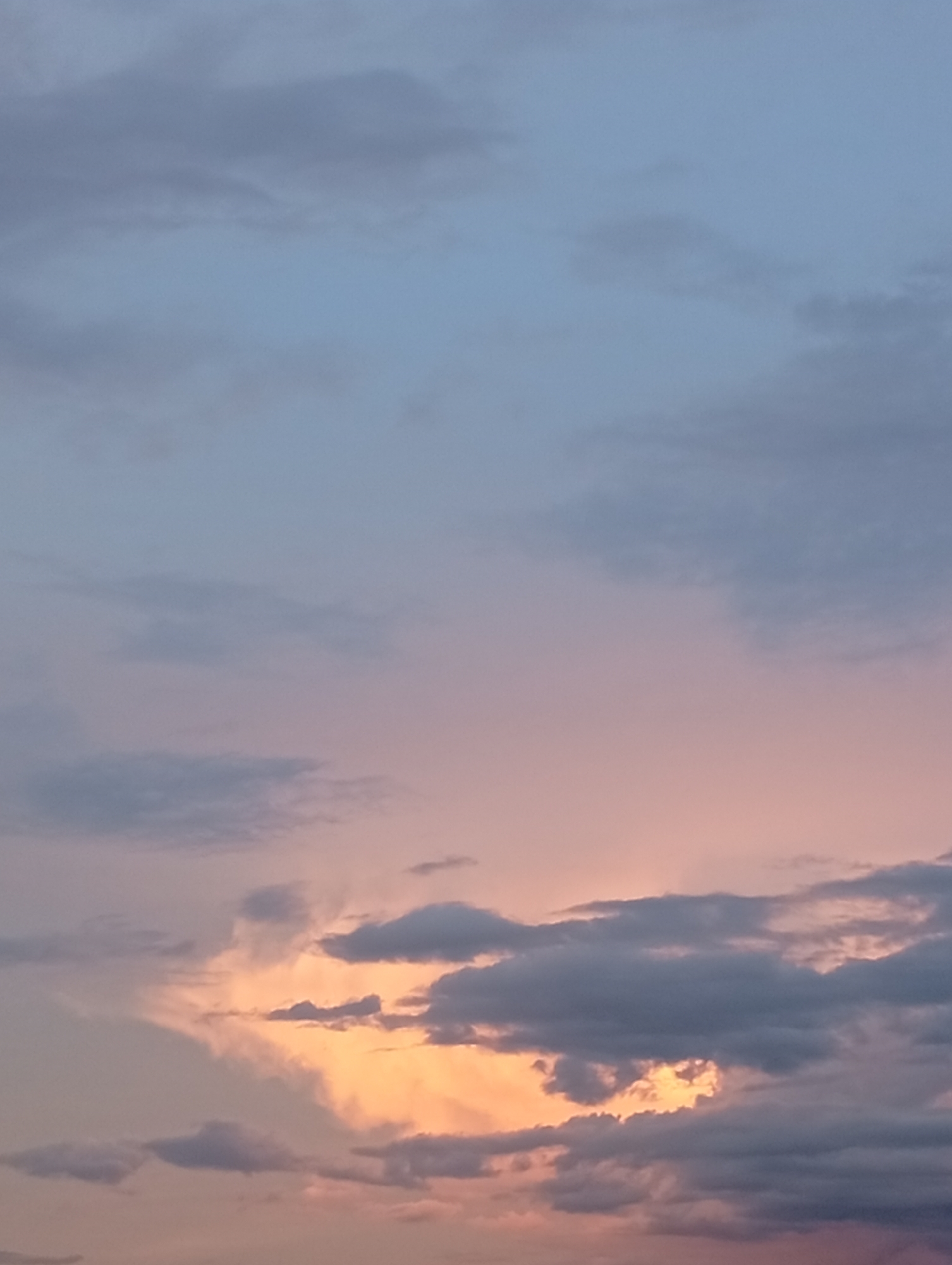 This media file displays a sunset sky with dramatic cloud formations. The upper portion of the image features a pale blue and soft lavender gradient, indicative of the fading daylight. Lower down, the clouds are illuminated by the setting sun, creating a vibrant display of orange, pink, and peachy hues that contrast with the darker, silhouetted clouds. These darker clouds are textured and varied, some appearing wispy and thin, while others are denser and more defined. The interplay of light and shadow across the clouds creates a sense of depth and movement. The overall atmosphere is serene and awe-inspiring, characteristic of a beautiful sunset. There are no people, objects, or discernible location cues beyond the sky itself. The time of day is clearly sunset. The weather appears calm, with the clouds suggesting a clear evening. There is no visible text in the image.