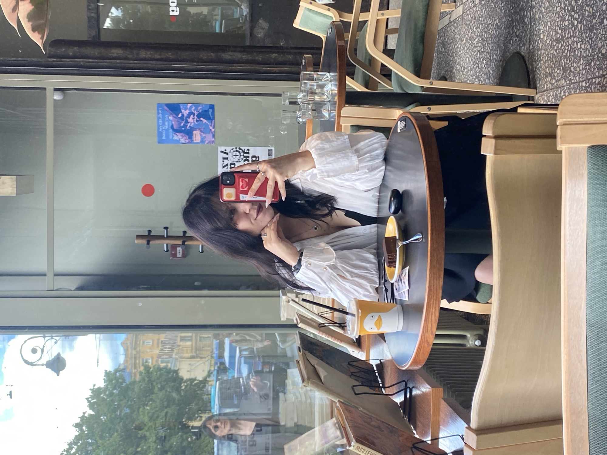 The image shows a woman taking a selfie in a cafe with large windows overlooking a city street. She is sitting at a round table with a slice of chocolate cake, a cup with a duck on it, and a phone in her hand. The cafe appears to be located in Saint Petersburg, Russia, as indicated by the Cyrillic text on the building visible through the window and the architectural style of the building in the background. The woman is wearing a white shirt with ruffles and a black dress. The cafe has wooden tables and chairs, and plants are visible. The overall scene suggests a casual cafe setting in an urban environment.