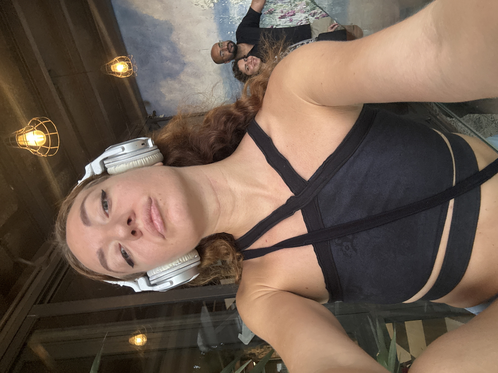 A woman with reddish-brown hair and headphones on her head is taking a selfie. She is wearing a black crop top and is positioned in the foreground of the image. In the background, a man and a woman are visible. The setting appears to be an indoor space with exposed wooden beams and decorative lighting fixtures overhead. A mural of clouds and possibly flowers is on the wall behind the people in the background. The floor has a checkered pattern. The overall impression is a casual, social setting.