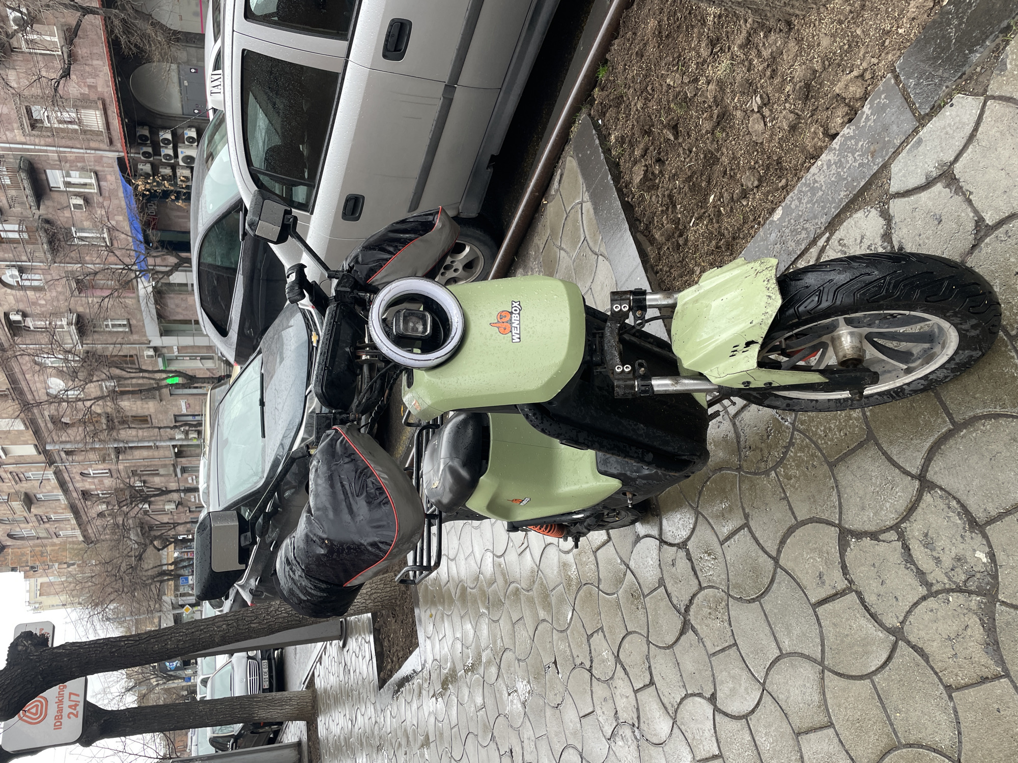 The image presents a forensic view of an urban street scene in Yerevan, Armenia, characterized by wet surfaces. In the immediate foreground, a light green "WENBOX" electric scooter is parked on a wet, patterned grey concrete sidewalk. The scooter features a circular LED headlight with an integrated camera, black handlebar muffs with red trim, a black seat, and a black storage box mounted on a rear rack; orange shock absorbers are visible. A patch of wet dirt with a metal rail on its edge is adjacent to the scooter on the right.
The mid-ground includes several parked vehicles on a wet asphalt street, notably a silver sedan with a "TAXI" sign on its roof, a dark grey hatchback, and a black Mercedes-Benz sedan bearing an Armenian license plate (01 OE 033). Bare-branched trees are positioned between the sidewalk and the street, one of which supports a white sign with "IDBanking.am" and "24/7" displayed in red. The background consists of multi-story, stone-faced buildings with classical architectural details, including arched windows and balconies. Multiple air conditioning units are externally mounted on the building facades. The sky is overcast, and no individuals are present in the frame.