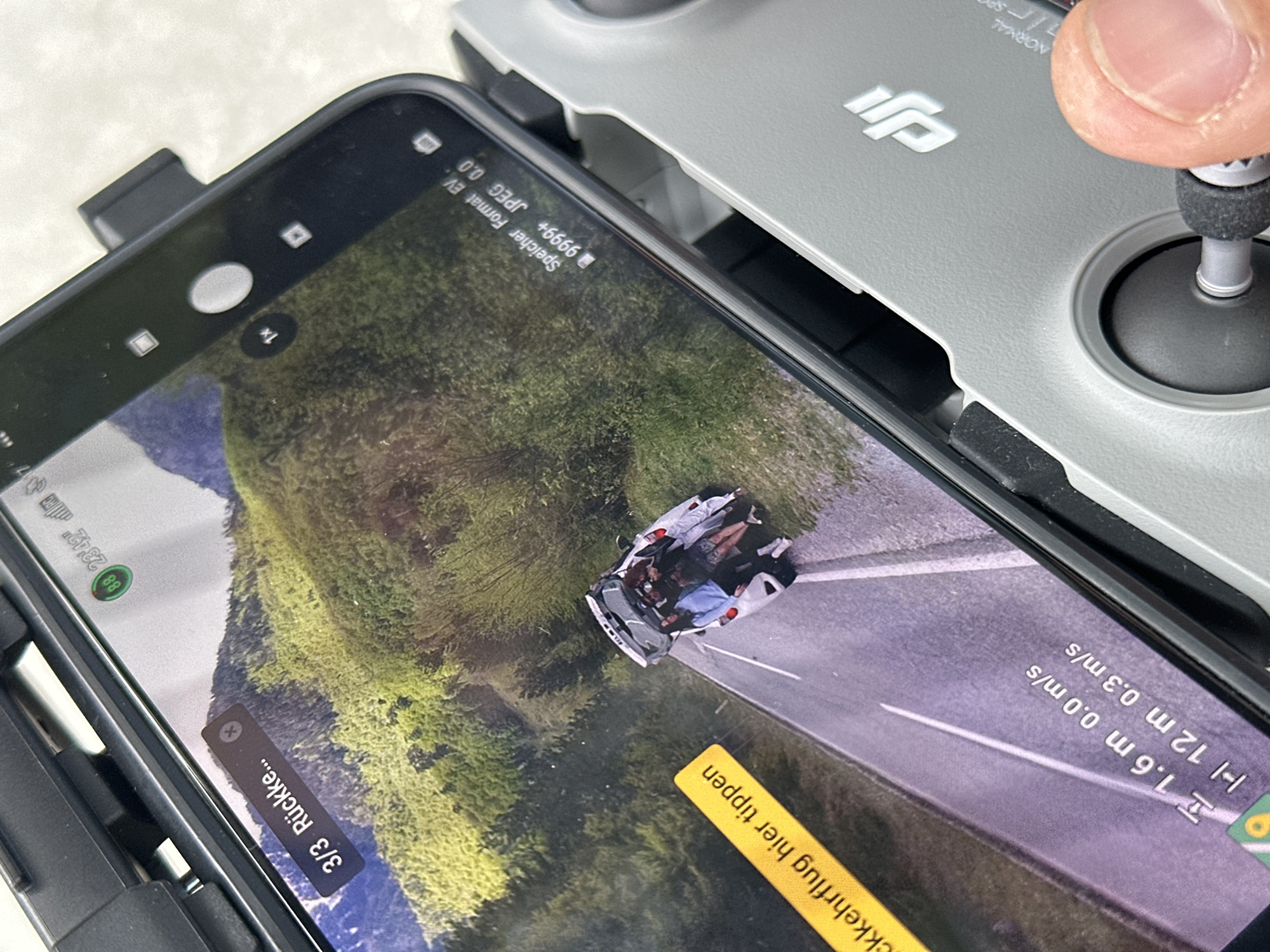 The image shows a phone screen displaying a drone's view of a convertible car on a road running near a green, mountainous terrain. The phone is resting on top of what appears to be a drone controller, and a human finger is visible near the controller's joystick. The phone's screen displays data such as "1.6 m 0.0 m/s" and "12 m 0.3 m/s", along with the text "3/3 Rückke...", which might be German for "return". Other text snippets include "ckkehrflug hier tippen" and "Speicher Format EV".
The location in the city of Bistrica, Montenegro is not evident in the image.