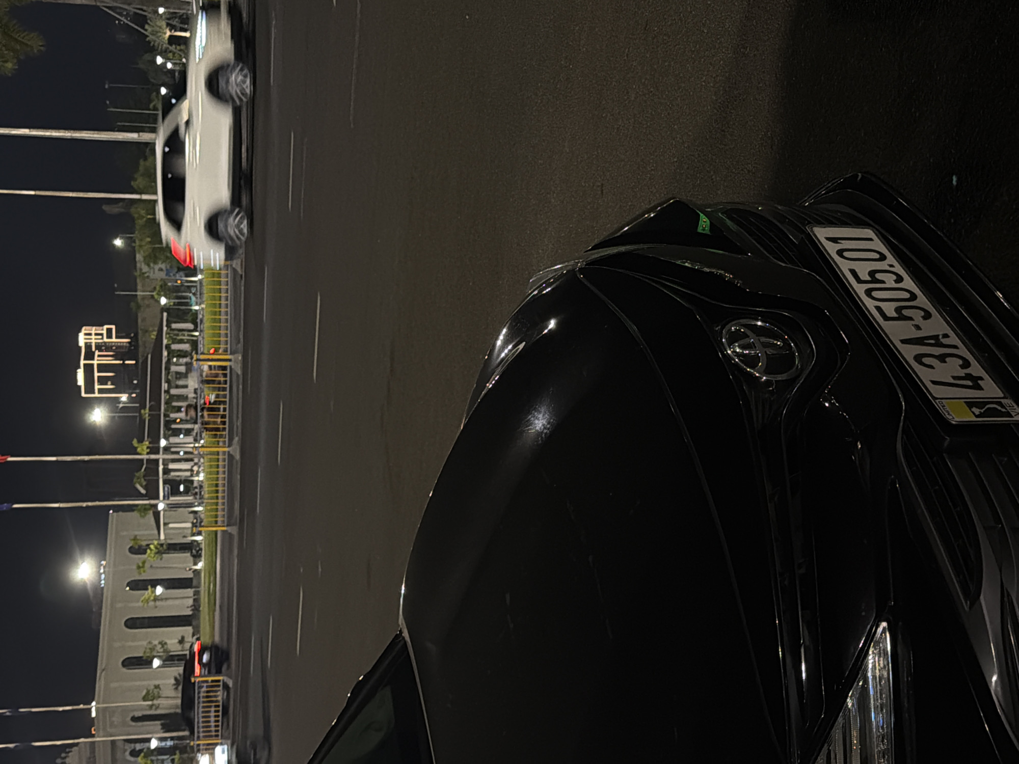 A black Toyota Camry is parked on the street at night, with its front end facing the camera. The car's license plate reads "43A-505.01". In the background, a white SUV is seen driving on the road, blurred due to motion. Beyond the road, there is a large, modern building with illuminated windows and a white facade. Flags on tall poles are also visible. The scene is lit by streetlights and the lights from the buildings. The image appears to be taken in Đà Nẵng, Vietnam, judging by the license plate format which is typical for the city.